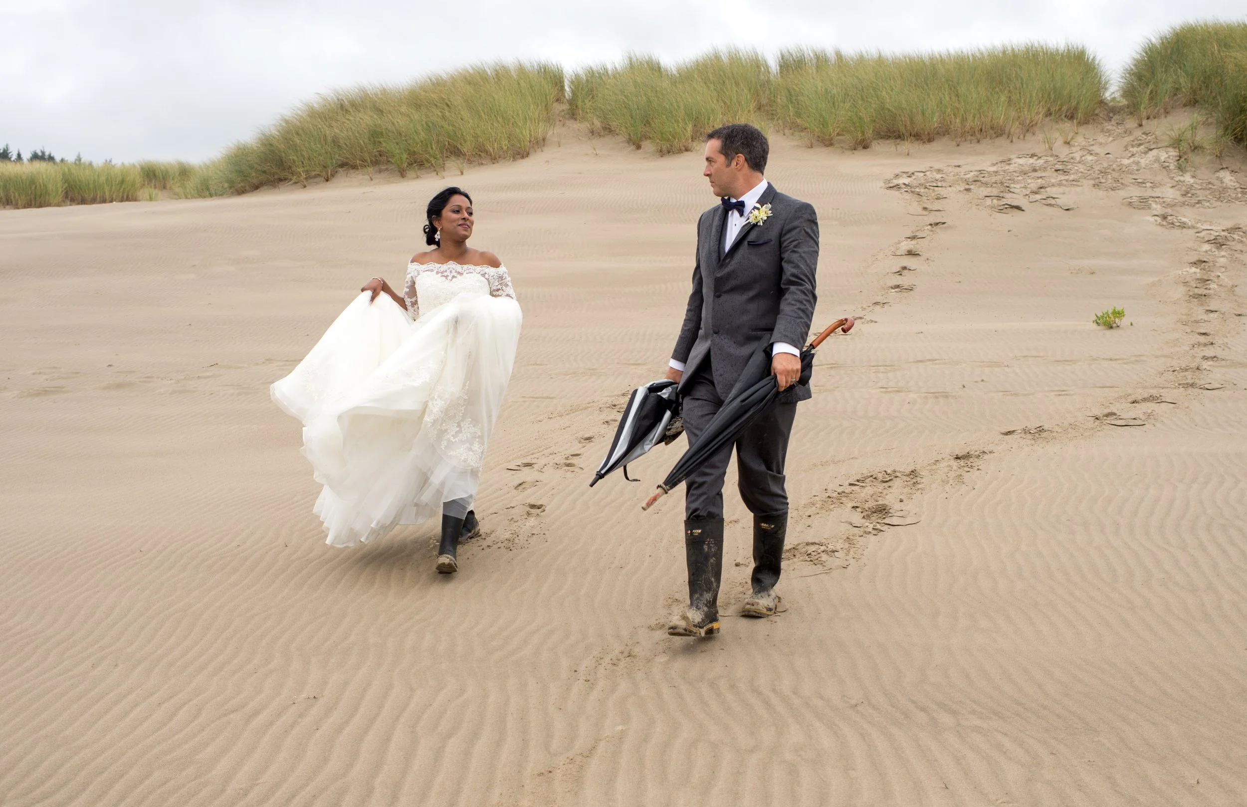 A bride in a white wedding dress and a groom in a gray suit walking on a sandy beach, holding umbrellas, with dune grass in the background under a cloudy sky.