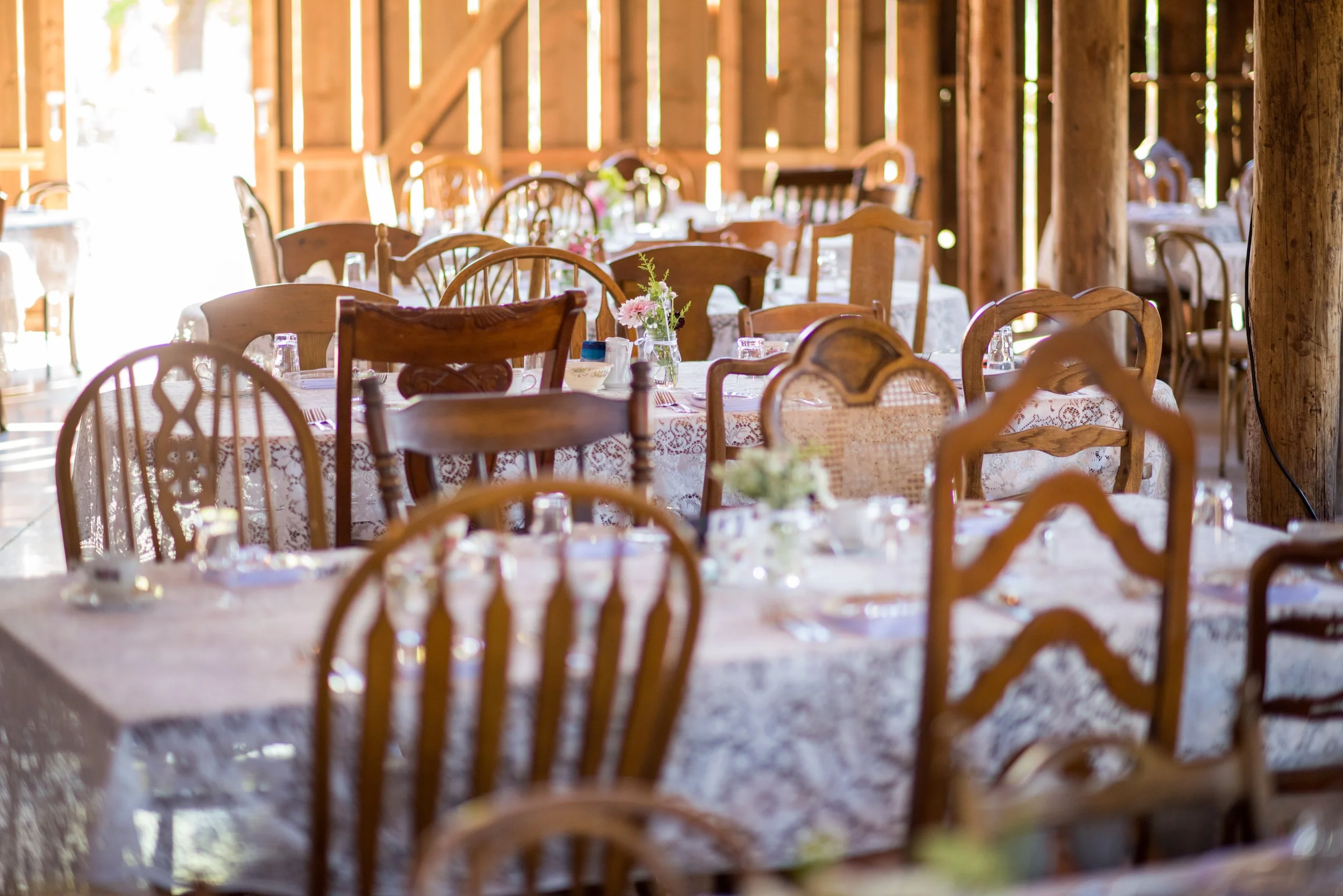 Wedding reception setup with round tables covered in lace tablecloths, decorated with floral centerpieces and glassware, inside a rustic barn with wooden beams and walls.