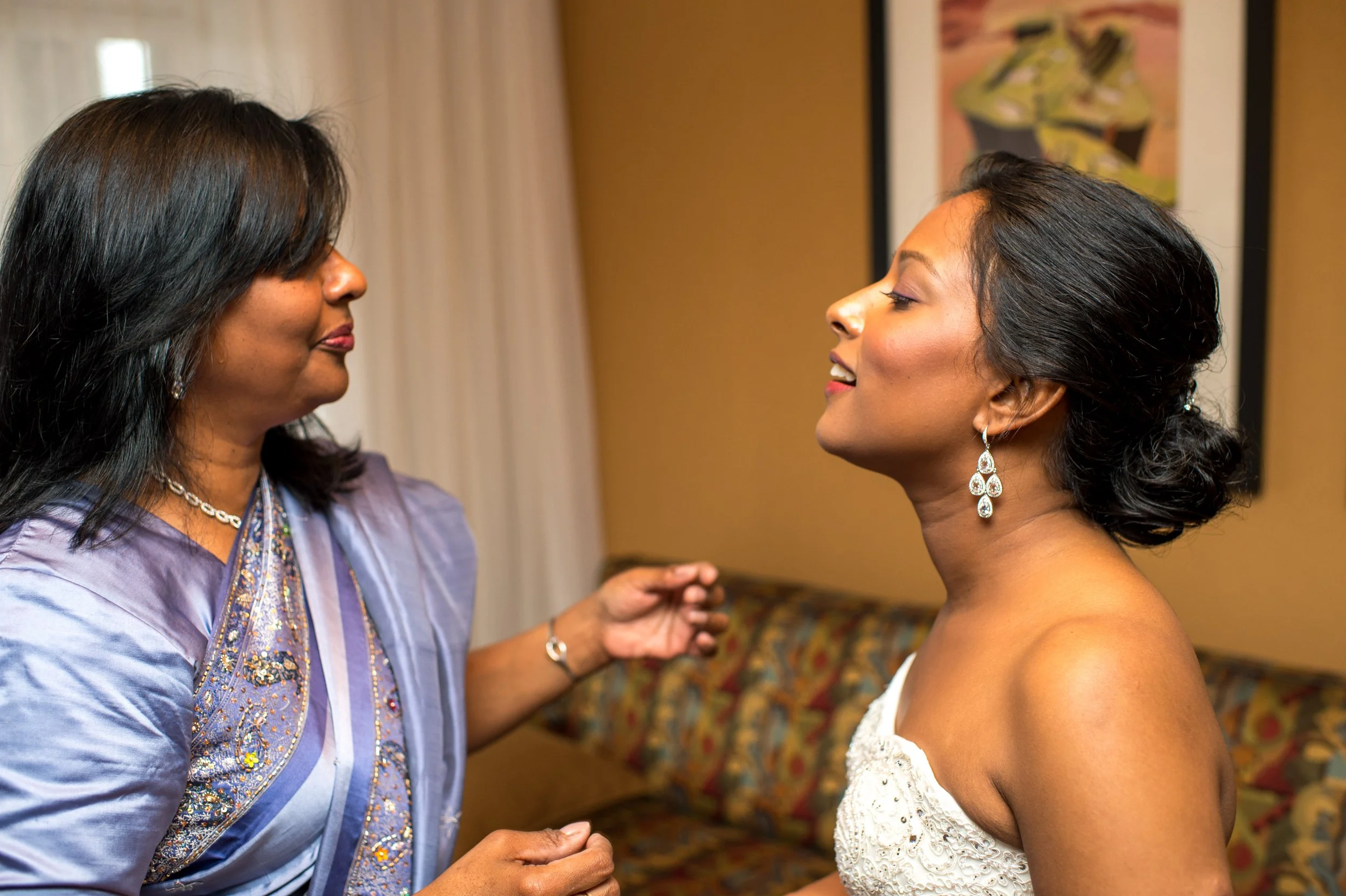 An older woman in a purple and blue embroidered dress is adjusting the earrings of a young woman in a white dress, both smiling softly. The background has a colorful floral pattern on the wall.