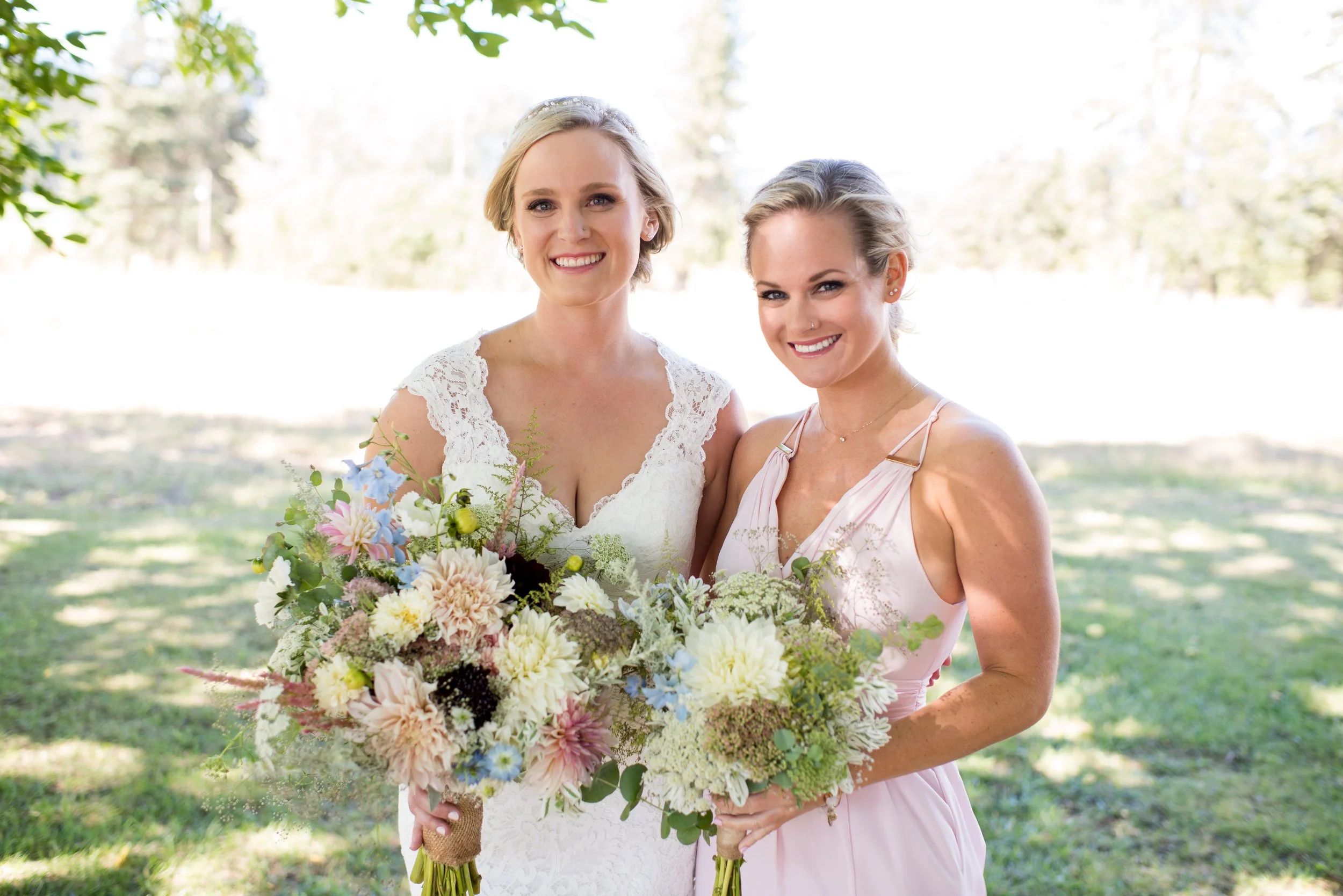 Two women smiling outdoors, one in a white lace dress holding a large bouquet of flowers, the other in a pink dress, standing next to each other on a sunny day with trees in the background.
