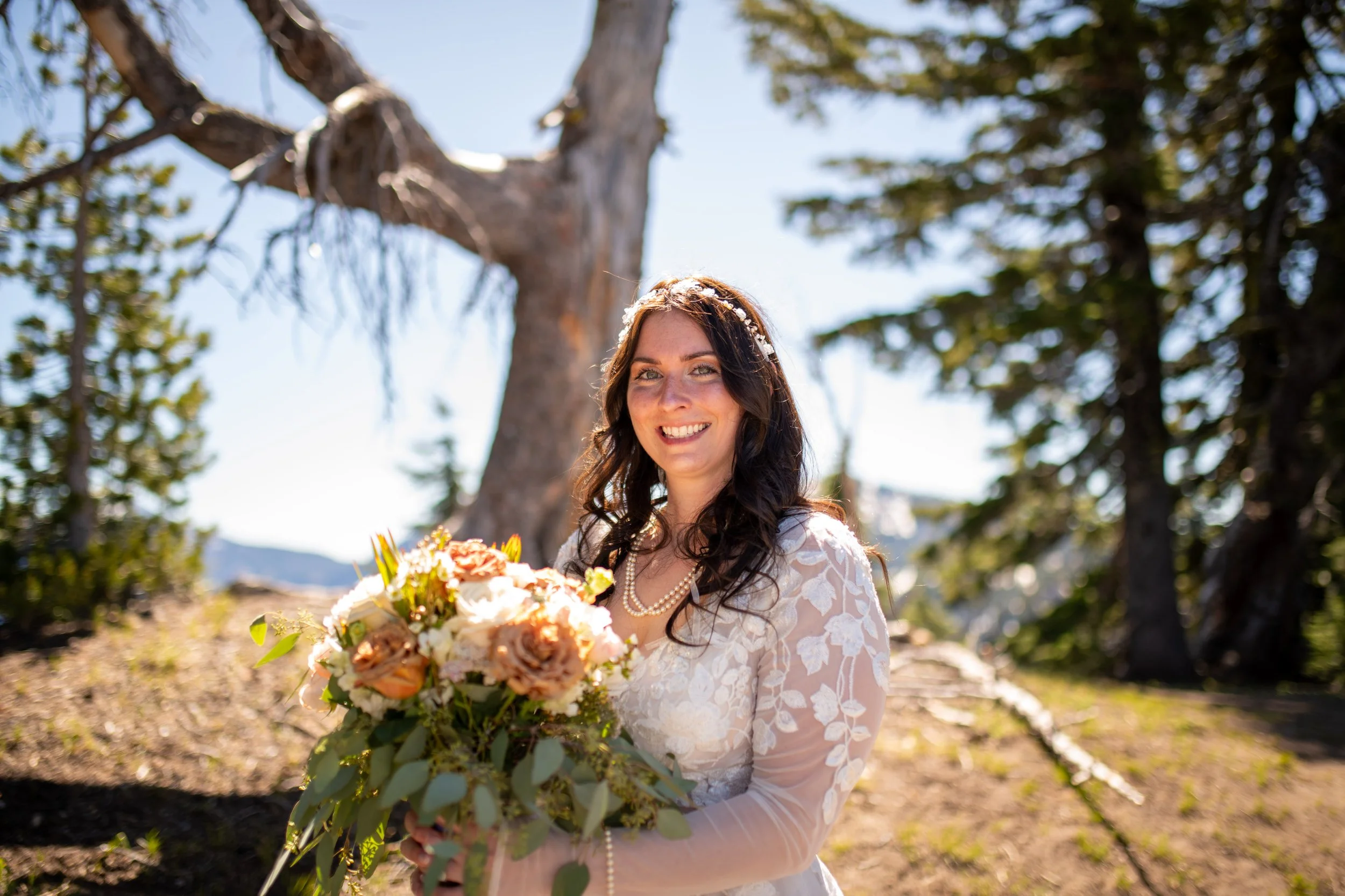 A woman smiling in a white lace dress holding a large bouquet of flowers outdoors in a wooded area with tall trees and a clear blue sky.