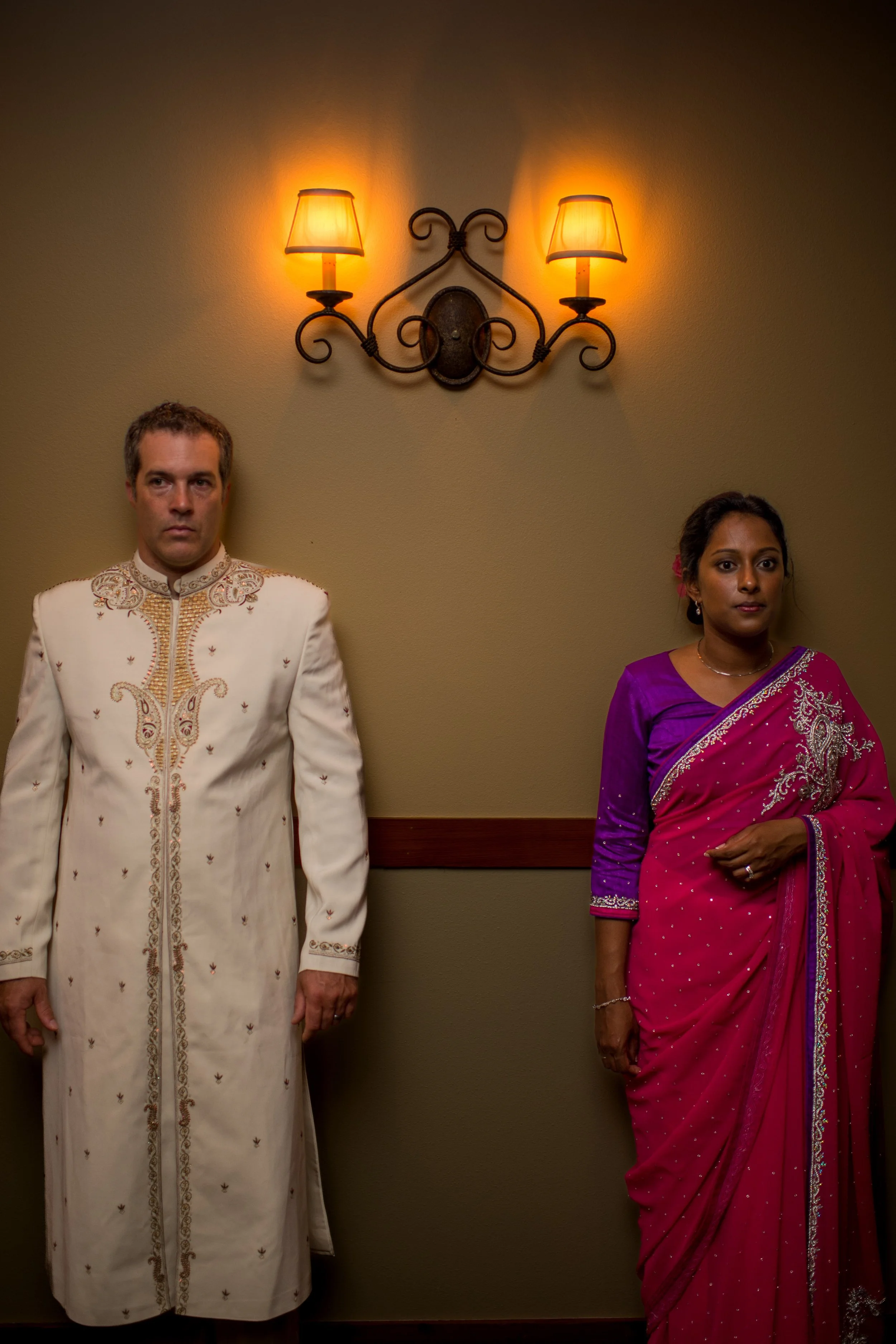 A man in traditional South Asian attire with embroidery details stands next to a woman in a vibrant pink sari with gold embellishments, both standing in a softly lit room with a wall-mounted candle-style light fixture.