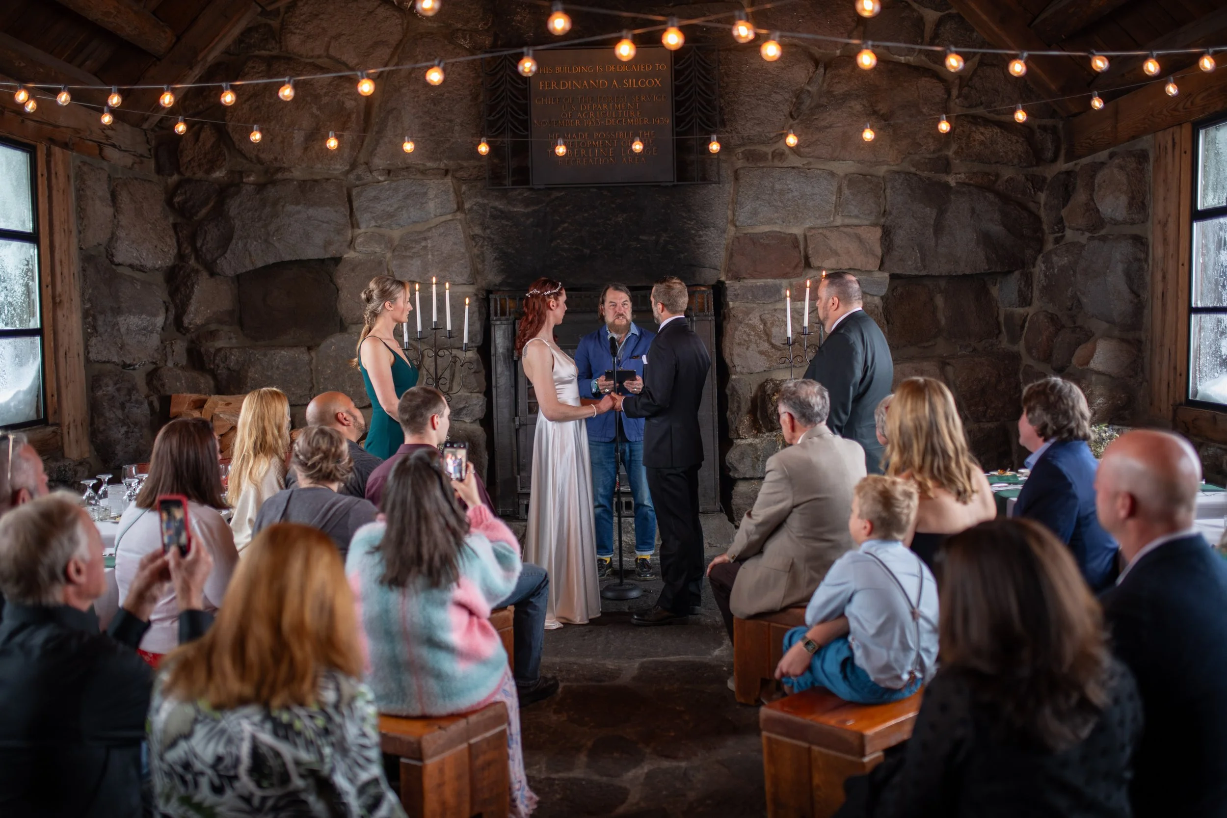 A wedding ceremony inside a rustic stone hall with a fireplace, decorated with string lights and candleholders. The bride and groom are exchanging vows, surrounded by guests seated at long tables.