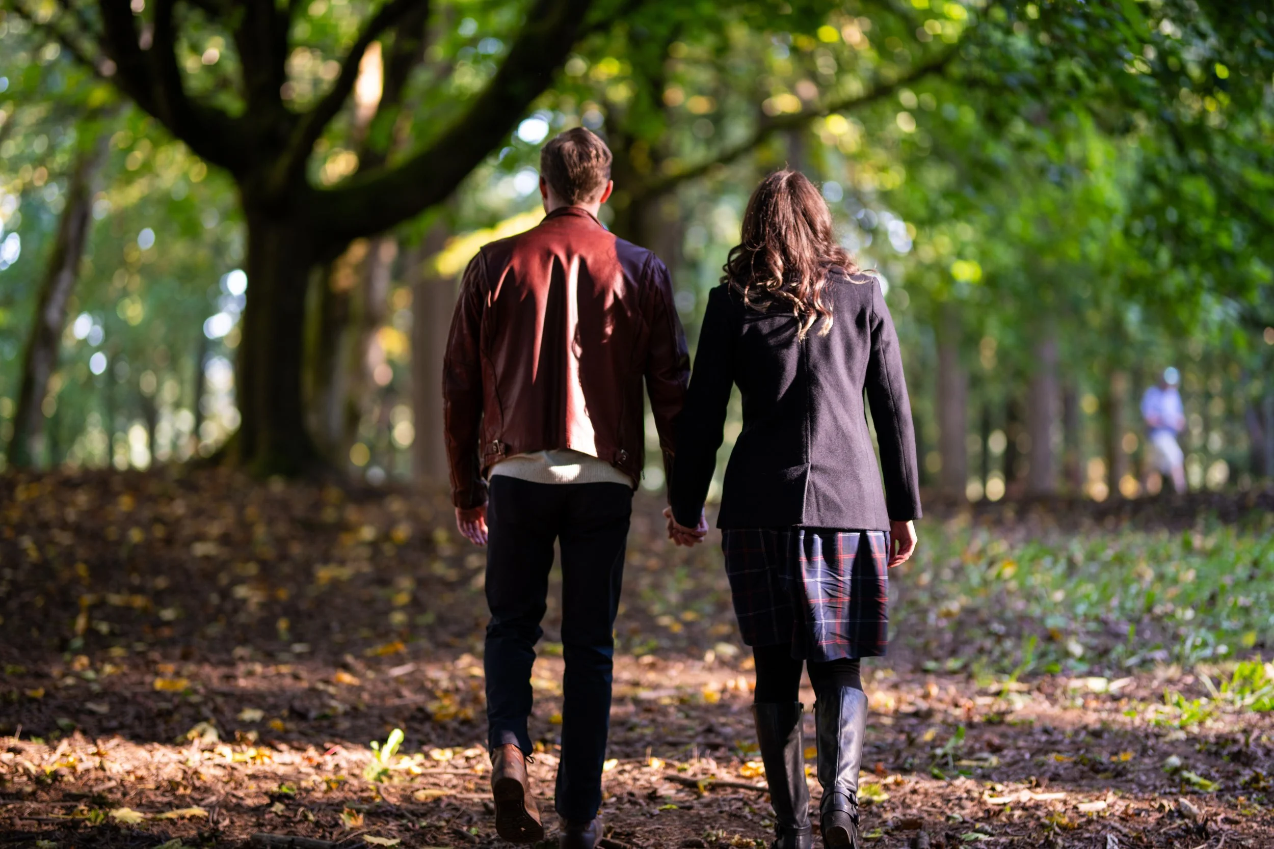 A couple walking hand-in-hand in a forest with green trees and fallen leaves on the ground.