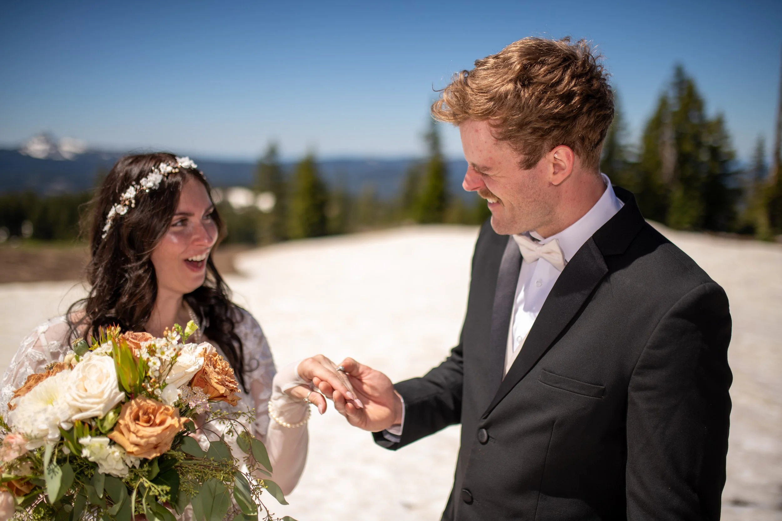 A bride and groom exchanging wedding rings outdoors on a sunny day with trees and mountains in the background.