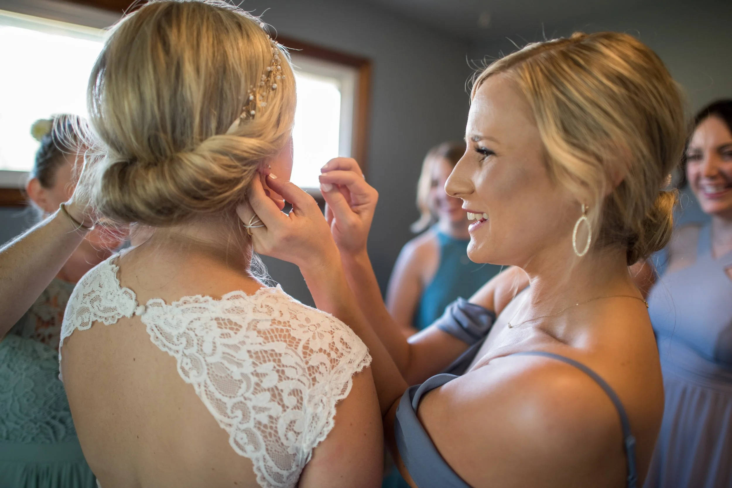 A woman in a blue dress is holding the face of another woman who is dressed in a white lace dress, in a joyful moment with other women smiling in the background.