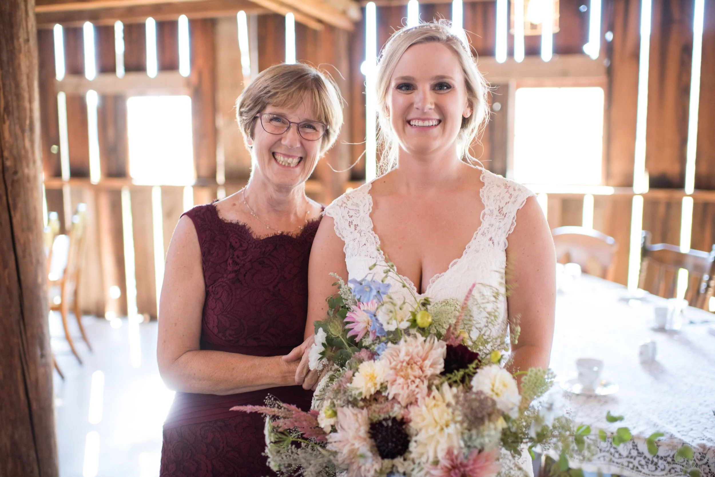 A bride holding a bouquet of flowers and another woman, both smiling, inside a rustic wooden venue with sunlight streaming through gaps in the wood.