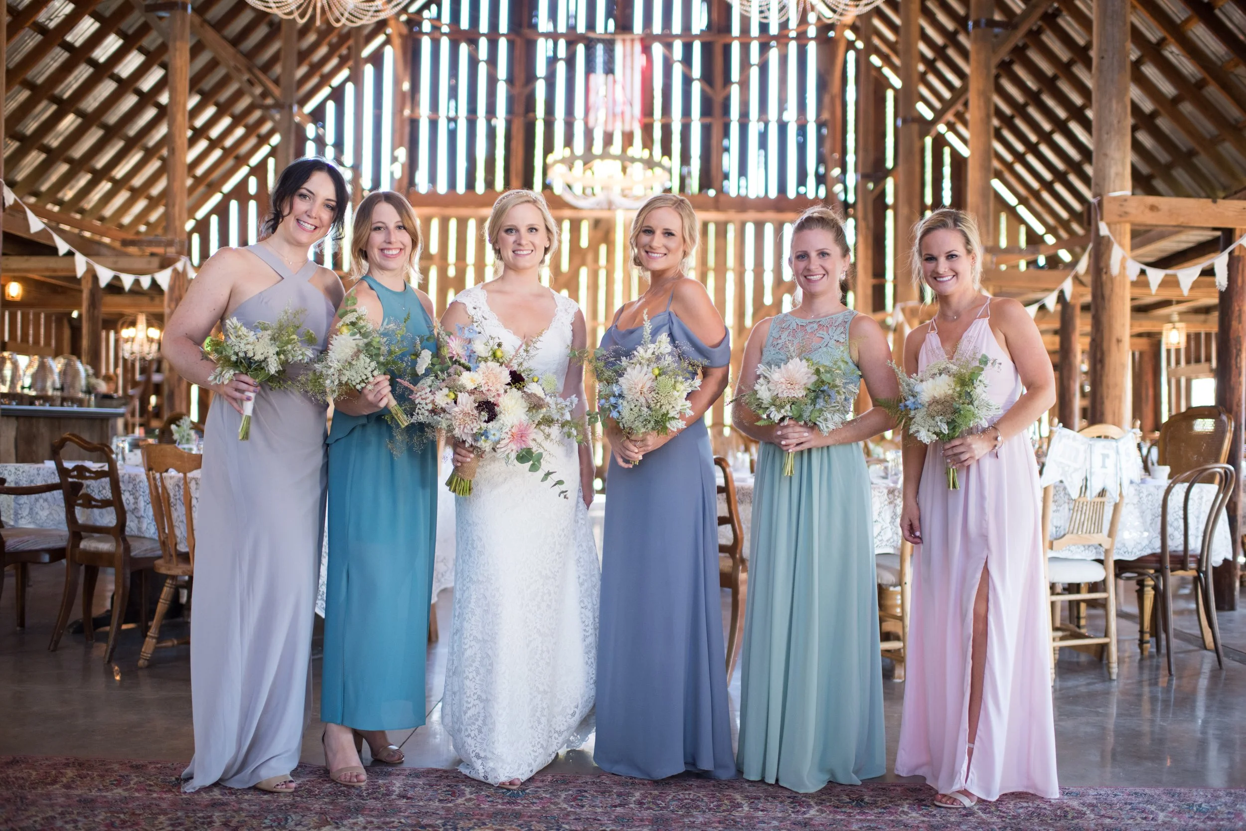 Six women in colorful dresses holding bouquets, standing inside a rustic wooden barn with high ceilings and string lights, during a wedding celebration.