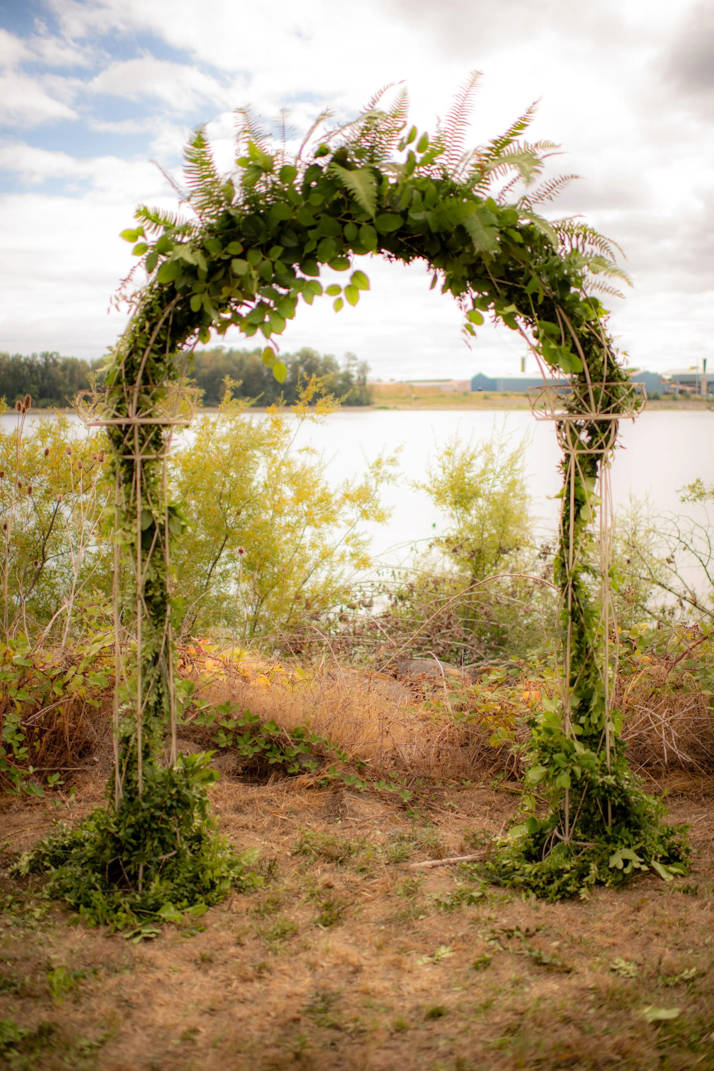 A decorative arch made of green leaves and ferns, standing near a body of water with a cloudy sky above.
