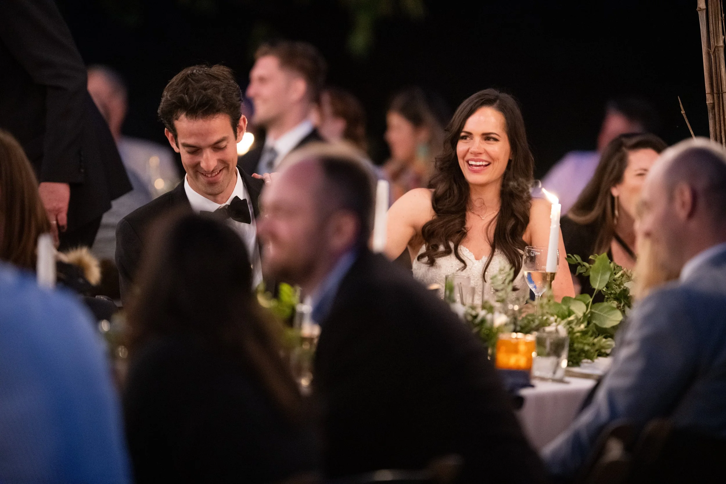 A group of people enjoying a wedding reception or party outdoors at night, with some people smiling and laughing, dressed in formal attire, and a table decorated with greenery and candles in the foreground.