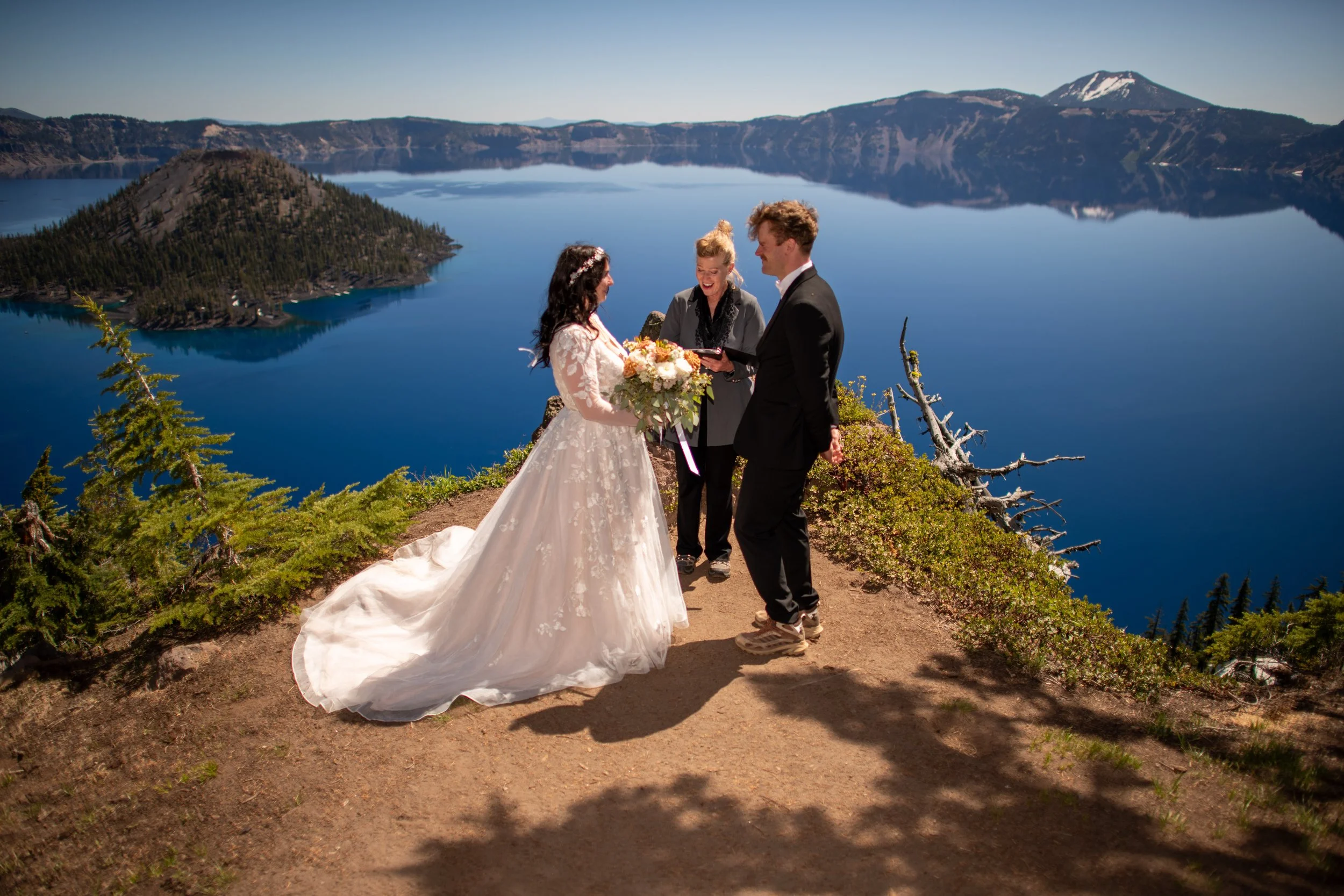 A wedding ceremony taking place outdoors on a dirt path by a large lake, with a mountainous landscape in the background. A bride in a white wedding dress and a groom in a dark suit are exchanging vows, accompanied by an officiant.