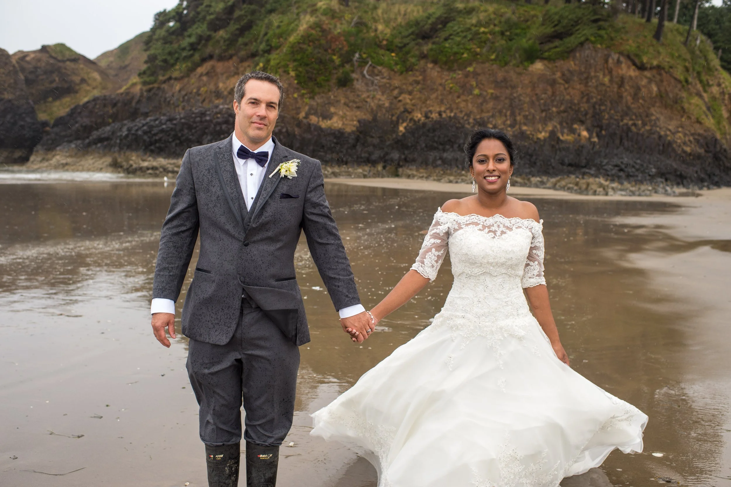 A bride and groom holding hands while walking along the beach near the water, with rocky cliffs and greenery in the background.