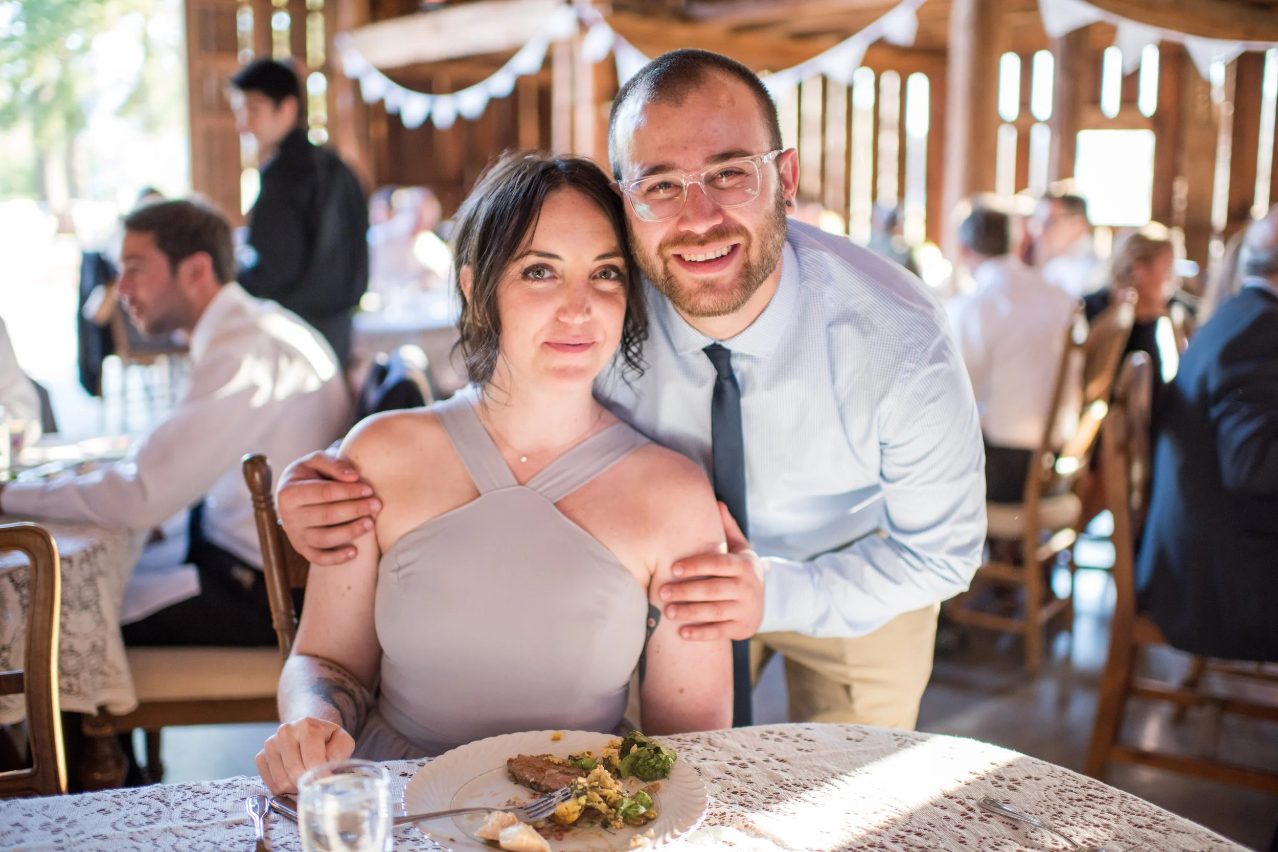 A happy couple sitting together at a formal event, smiling at the camera. The woman has dark hair and tattoos on her arm, wearing a light-colored dress. The man has a beard, glasses, and is dressed in a collared shirt with a tie. They are seated at a