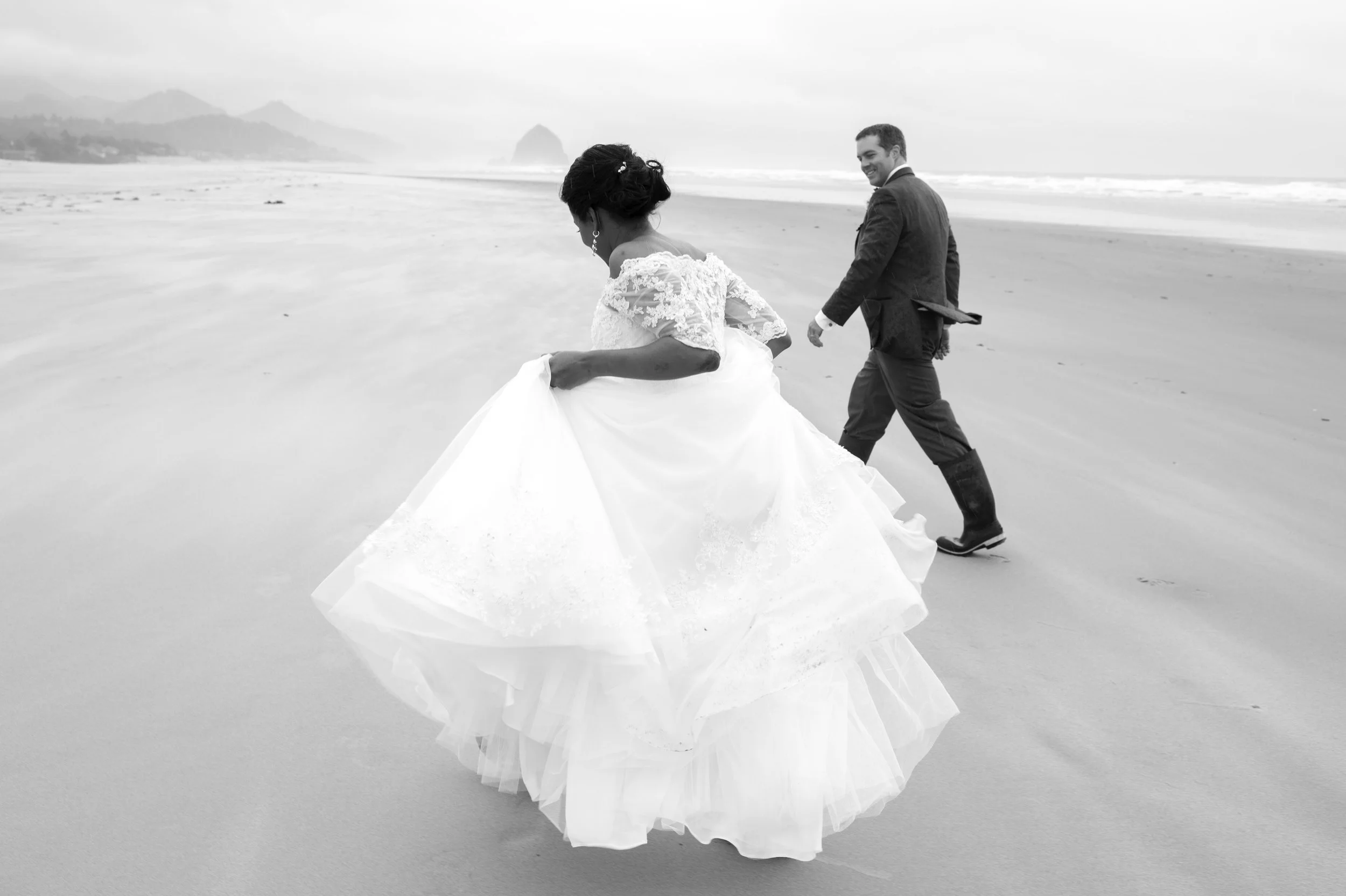 A bride in a wedding dress and a groom in a suit walk hand in hand on a beach with waves and a distant rocky island in the background.