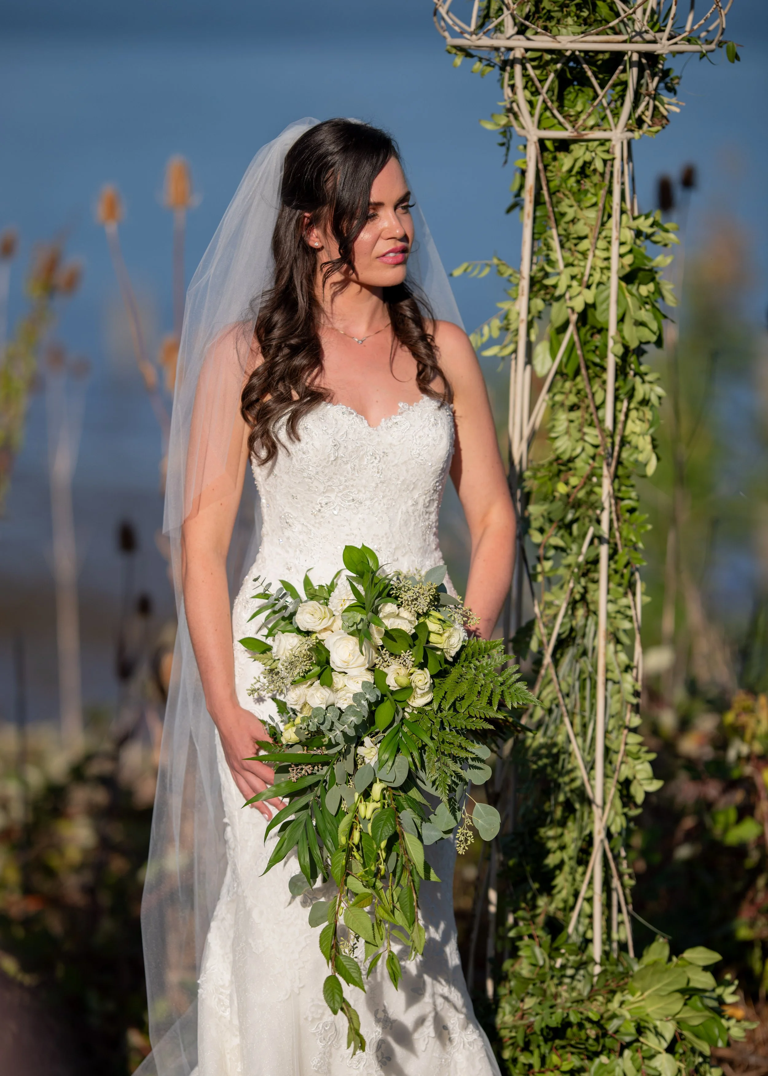 A bride in a white lace wedding dress with a veil and dark wavy hair holding a bouquet of white roses and greenery, standing outdoors near a decorative metal structure with green plants, against a background of blue sky and water.