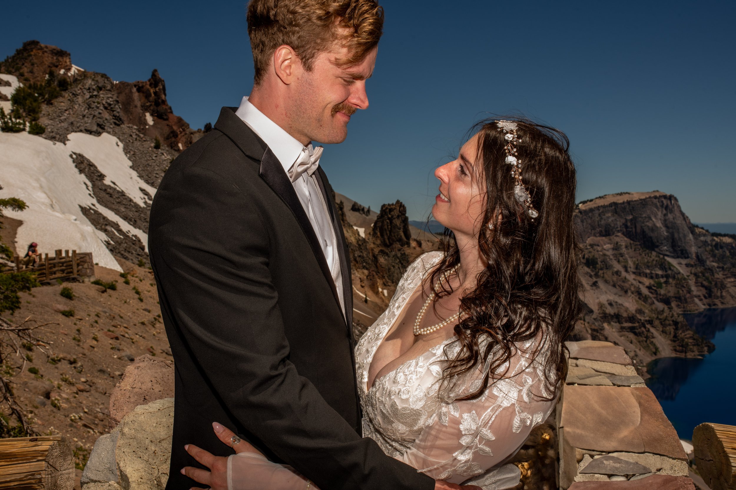 A bride and groom gazing at each other outdoors in a mountainous landscape with patches of snow, rocky cliffs, and clear blue sky, dressed in wedding attire.