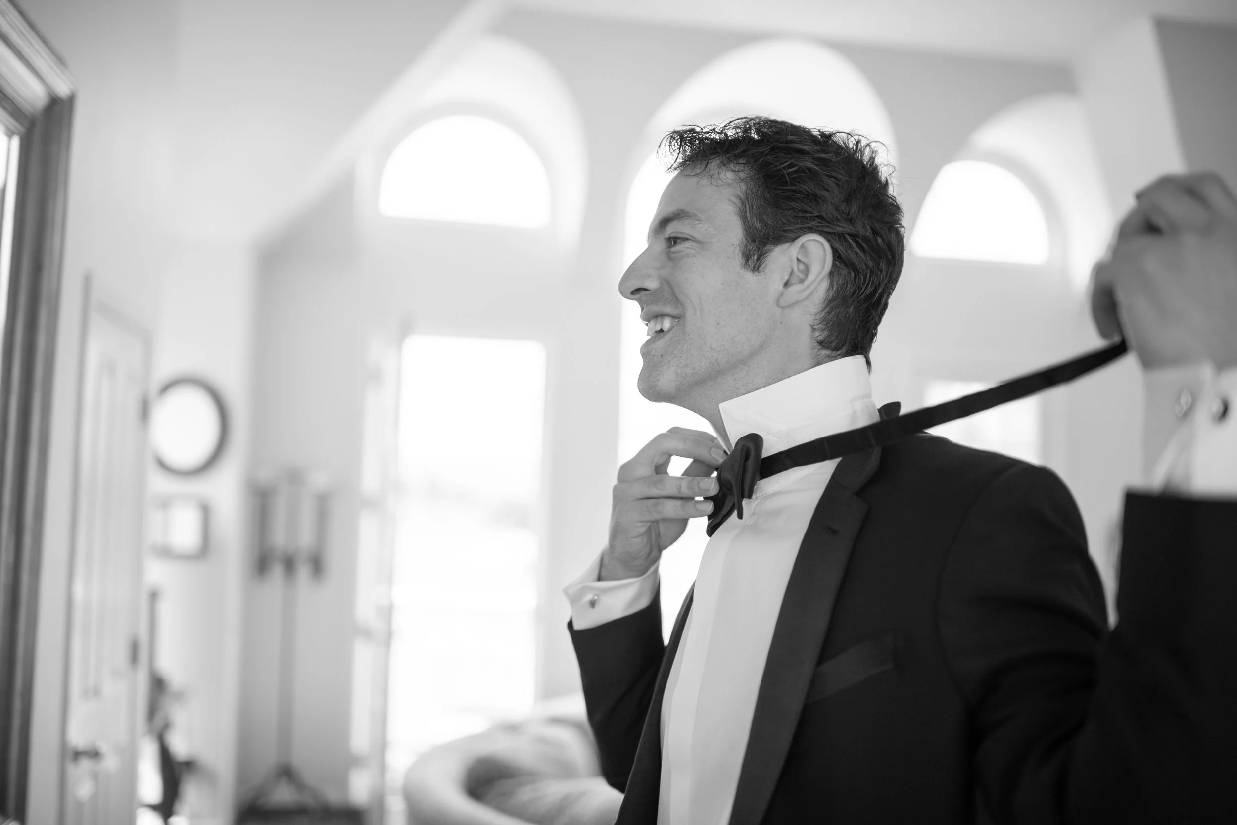 A man in formal attire, adjusting his bow tie, standing in a bright room with large arched windows.