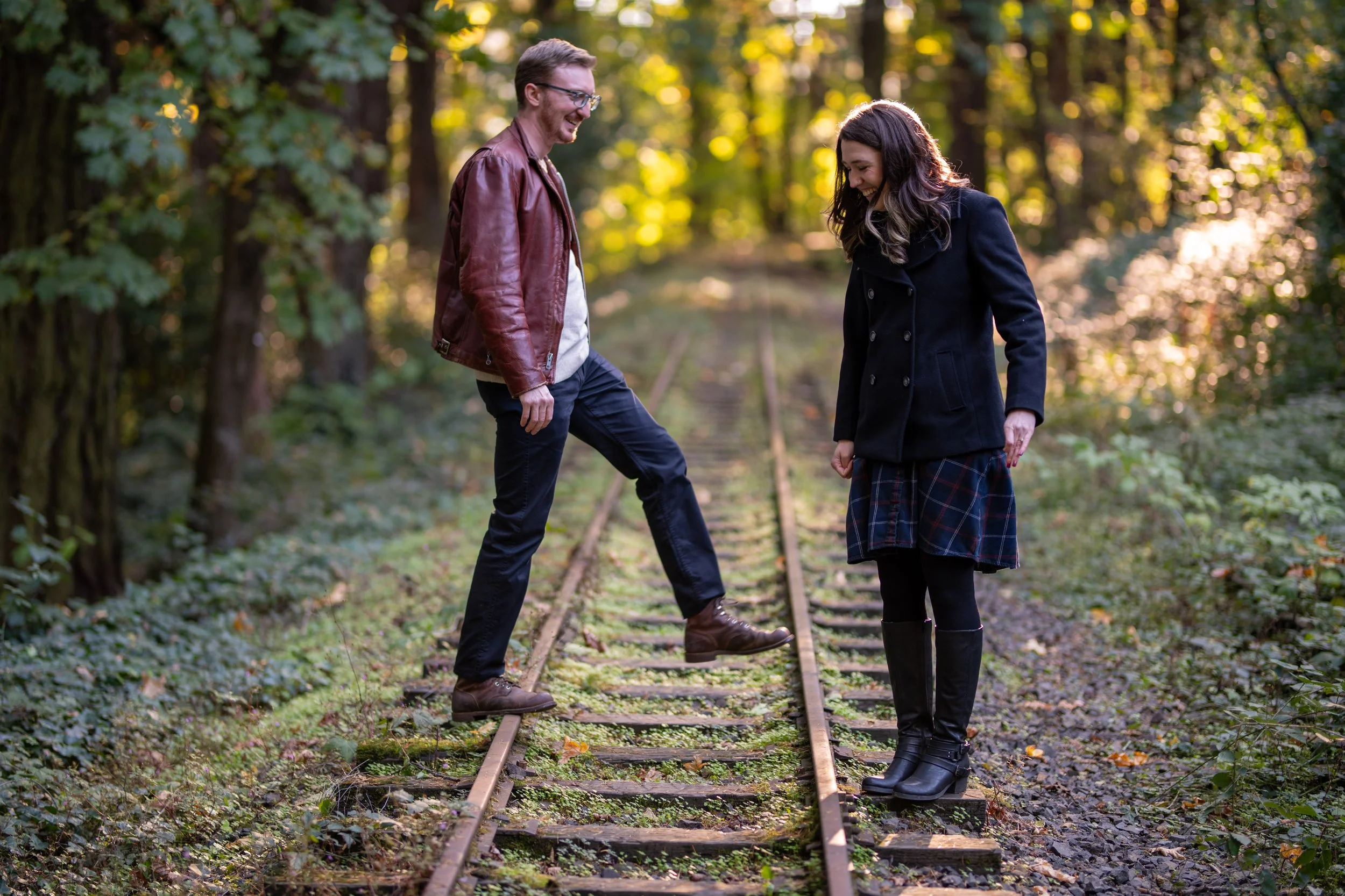 A man and woman standing on an old train track in a wooded area, smiling and laughing together, with sunlight filtering through the trees.