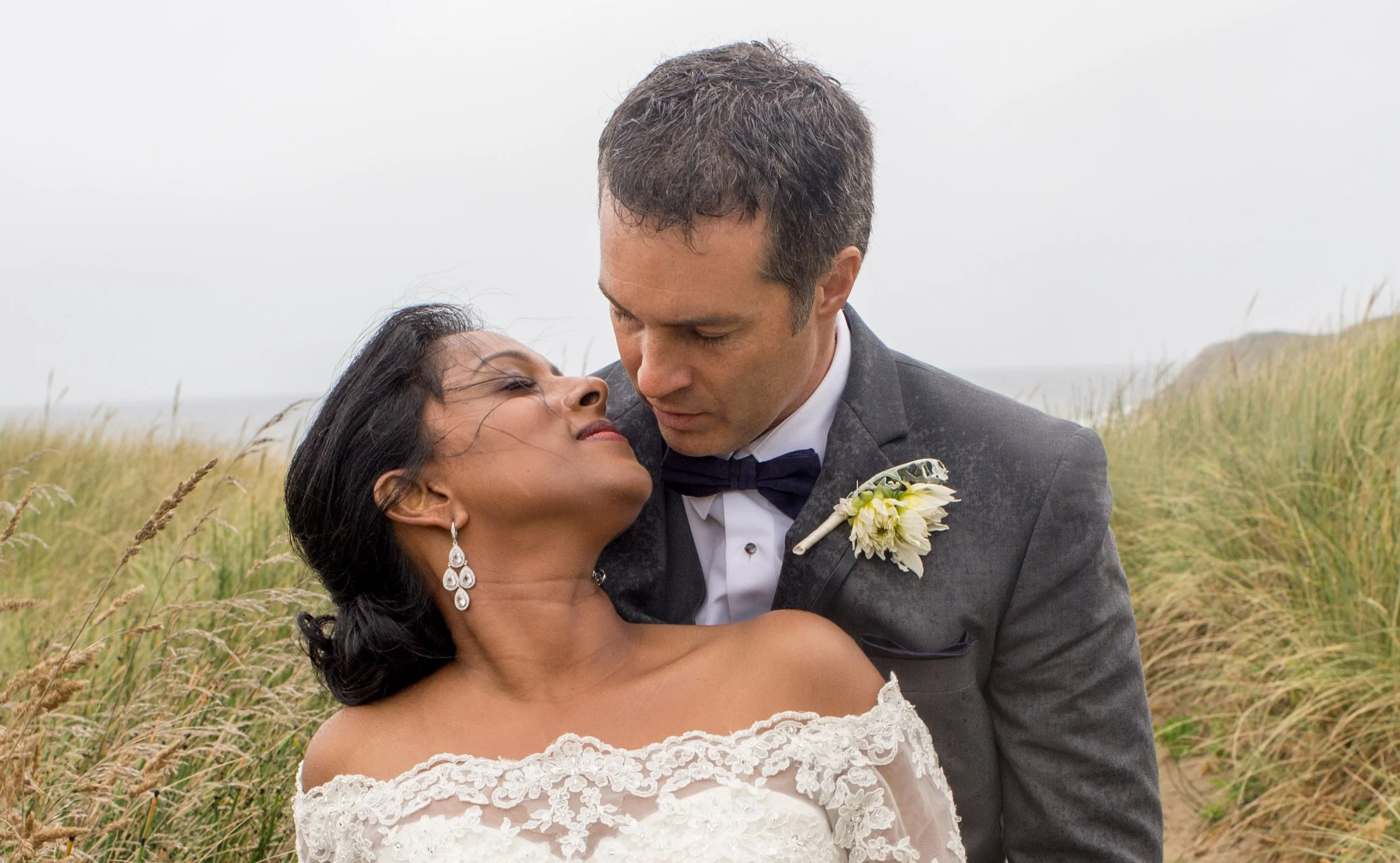 A bride and groom embrace outdoors among tall grass, with the bride looking up at the groom and the groom gazing down at her, both dressed in wedding attire.