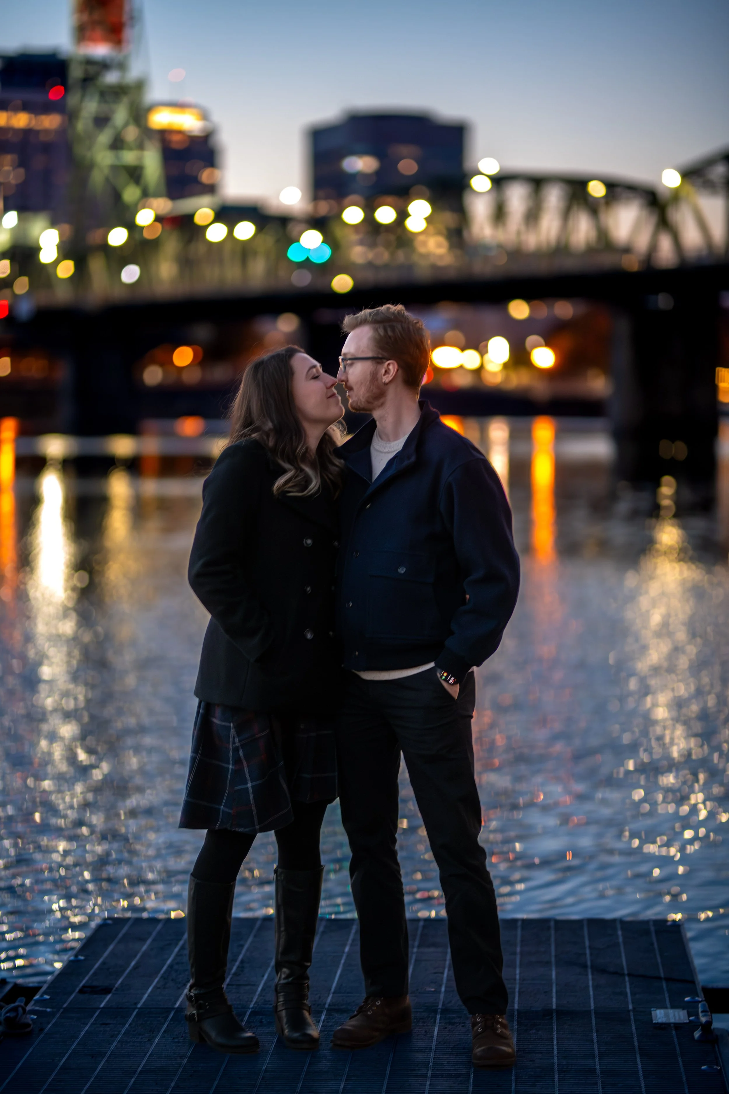 A couple standing close and touching foreheads on a dock by the water during sunset, with blurred city skyline and lights in the background.