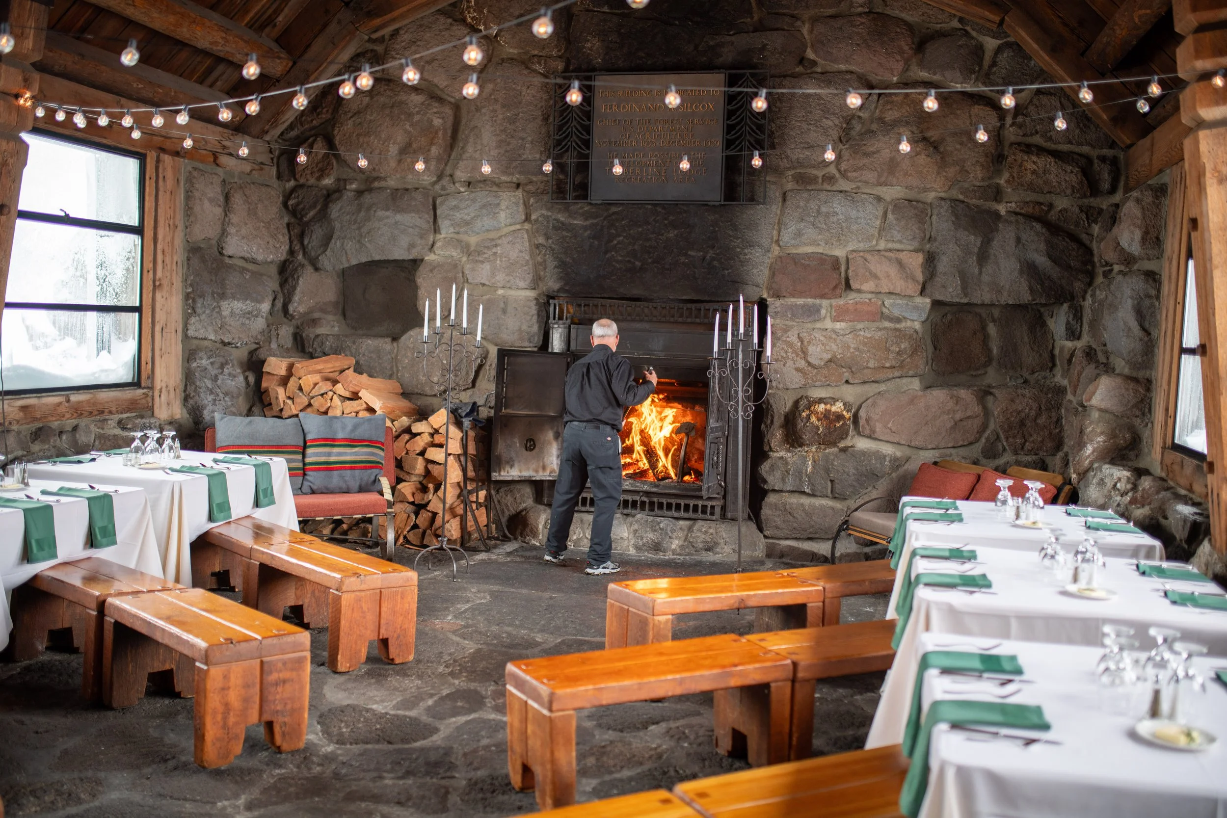 A cozy dining area inside a rustic stone building featuring a large fireplace with logs in the corner and string lights hanging from the ceiling. Tables are set with white tablecloths, green napkins, and glassware. A person is tending to the fire in 