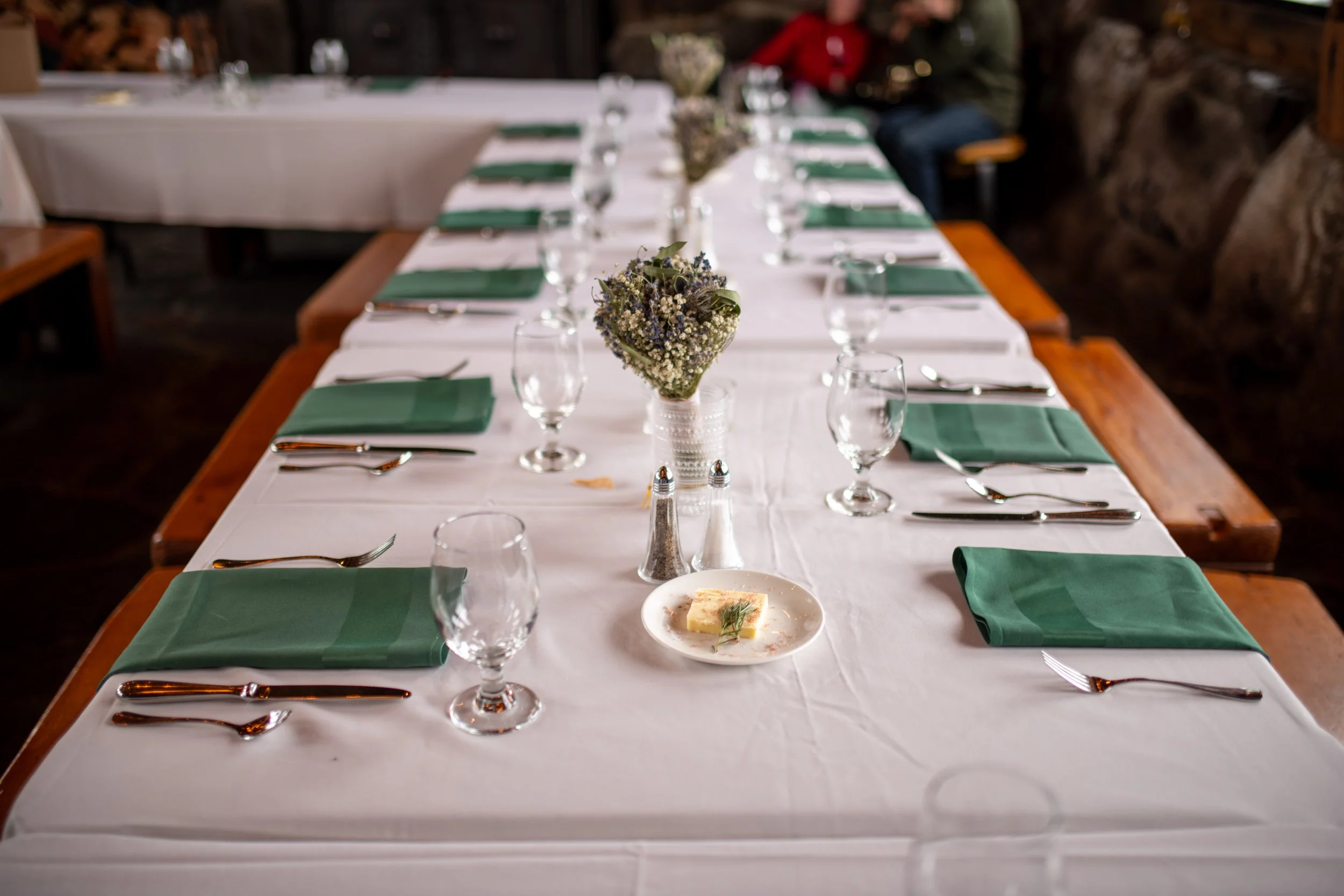 Long dining table with white tablecloth, green napkins, wine glasses, and silverware, decorated with a flower arrangement in a clear vase, in a rustic setting with rocks and wood in the background.