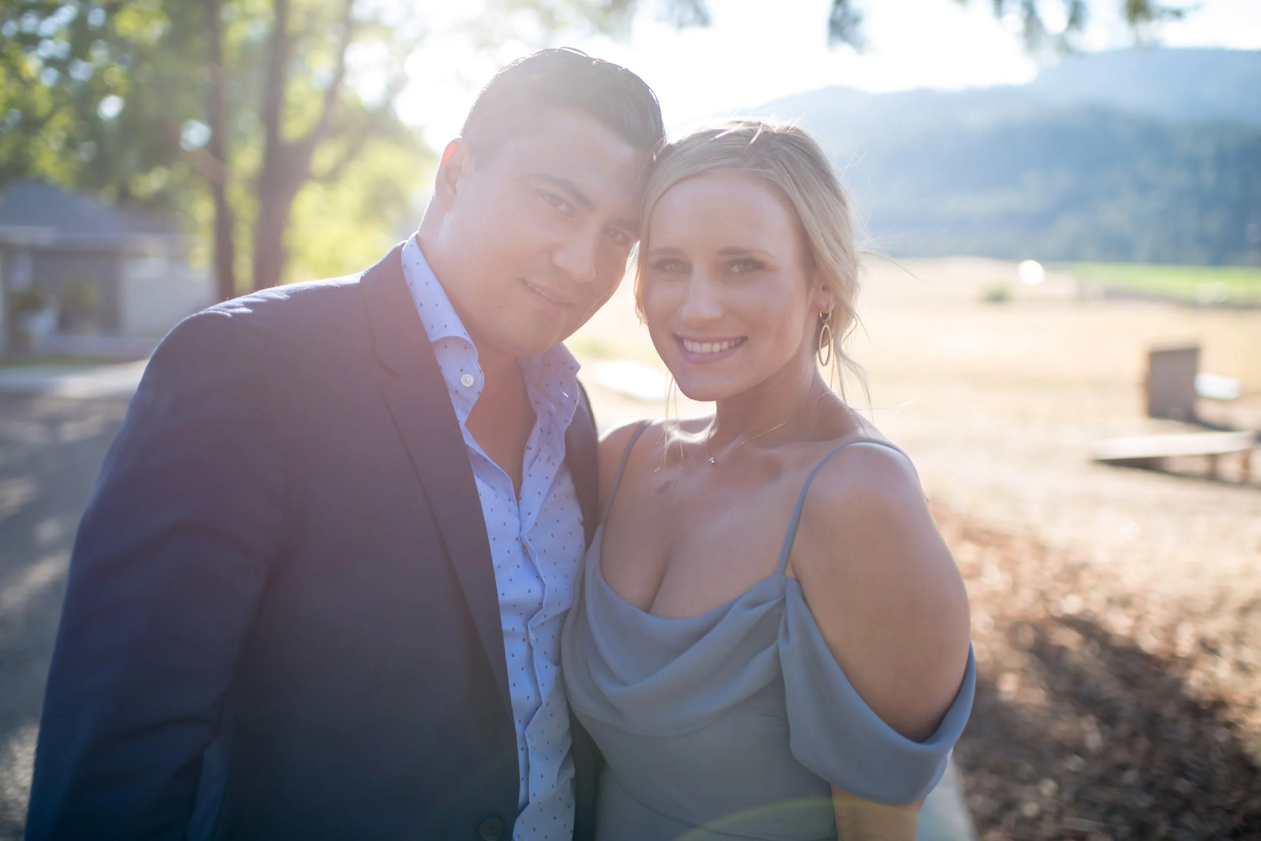 A smiling couple taking a selfie outdoors on a sunny day with trees and a mountain in the background.
