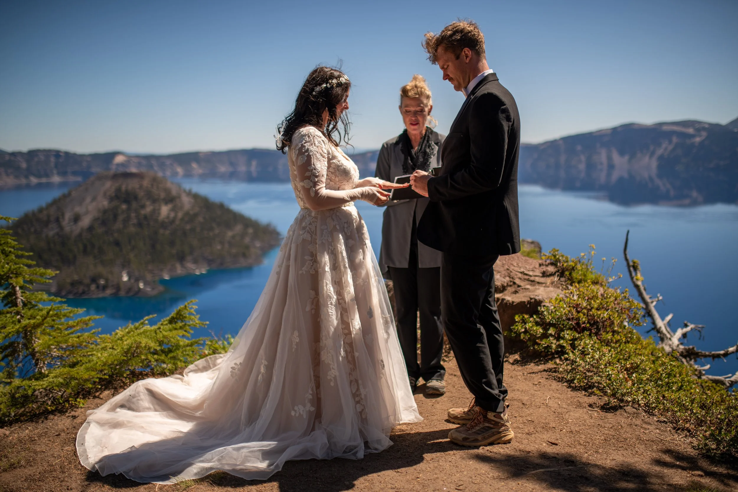 A couple getting married on a mountain overlook with a lake and mountains in the background, the bride in a lace dress and the groom in a tuxedo.