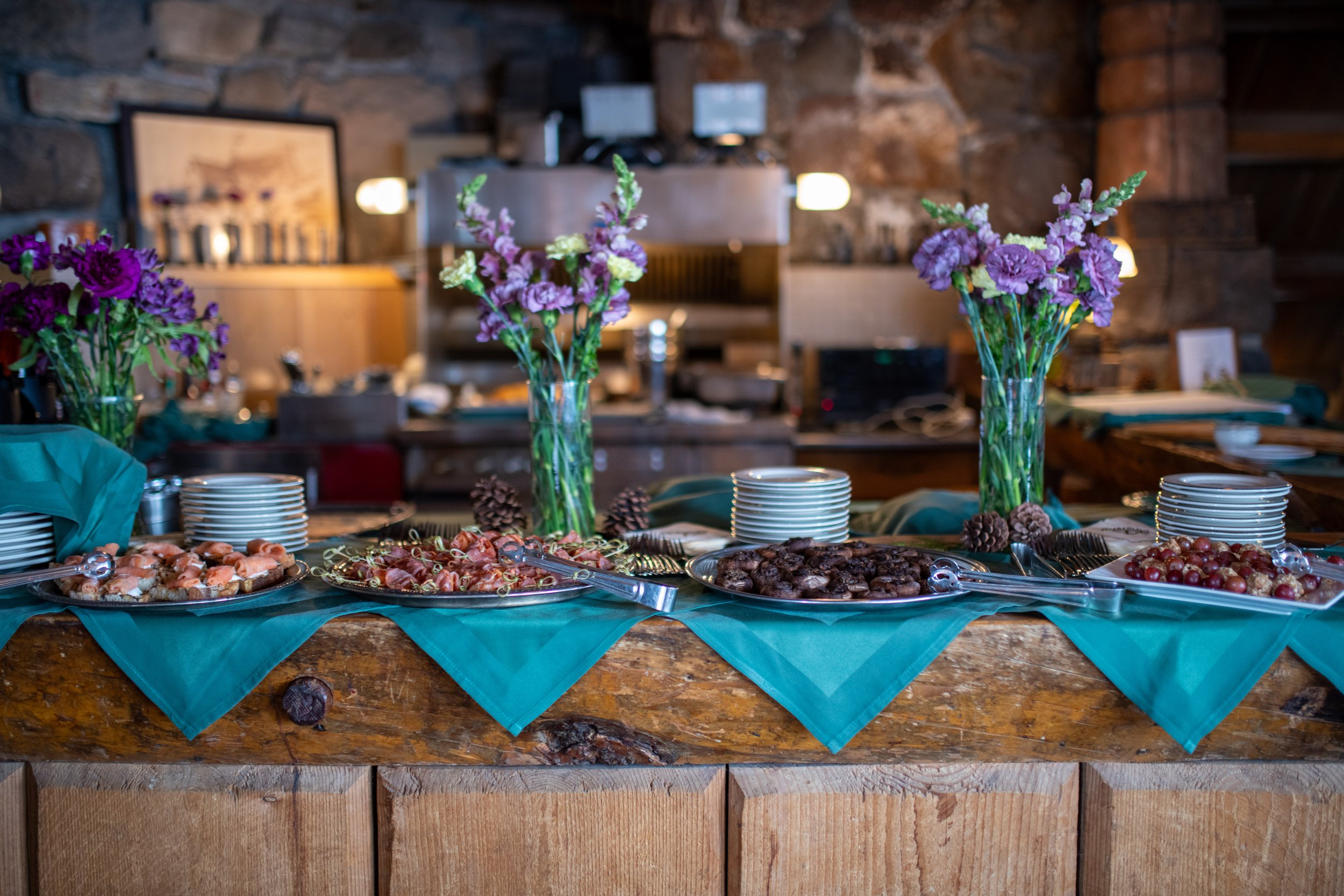 A buffet table with various snacks and refreshments, decorated with purple flowers in tall vases, set in a rustic setting with a stone wall background.