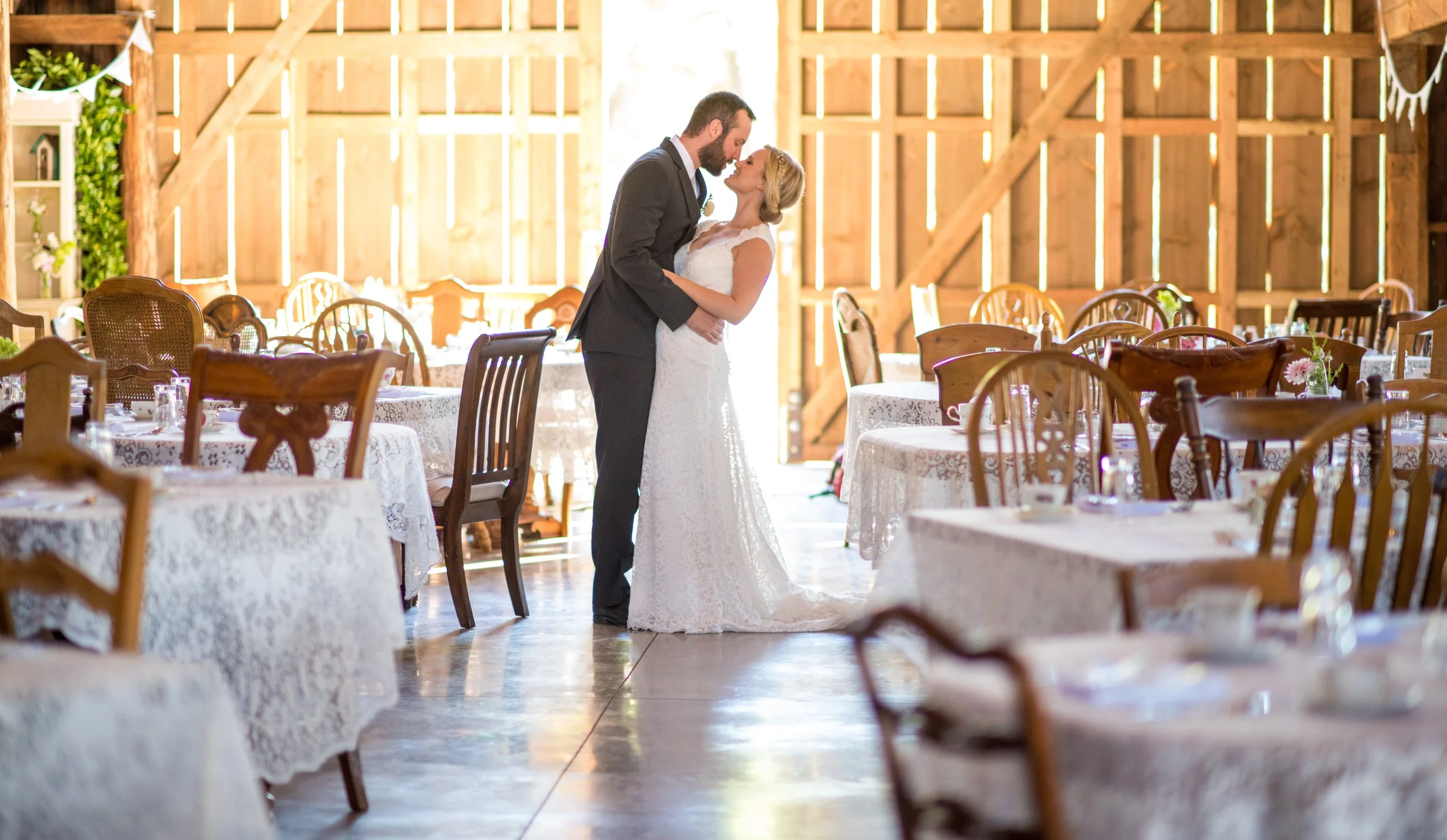 A bride and groom sharing a kiss inside a rustic barn decorated for wedding reception. The bride wears a white lace wedding gown, and the groom is in a dark suit. The barn has wooden walls and tables with white lace tablecloths.