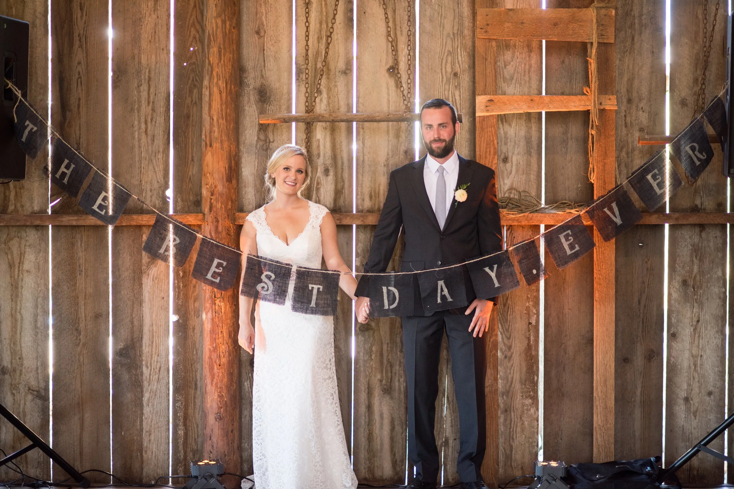 A bride and groom stand hand in hand in front of a rustic wooden backdrop with vertical slats and chains, during their wedding celebration.