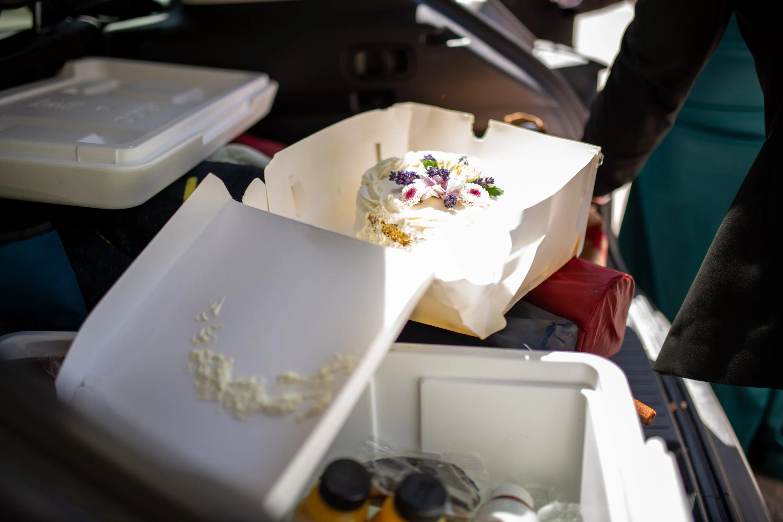 A partially eaten birthday cake with white frosting and purple and pink flowers on top is inside a white takeout box. The box is on a table at an event, with a person in dark clothing reaching towards the cake. There are other objects and party suppl