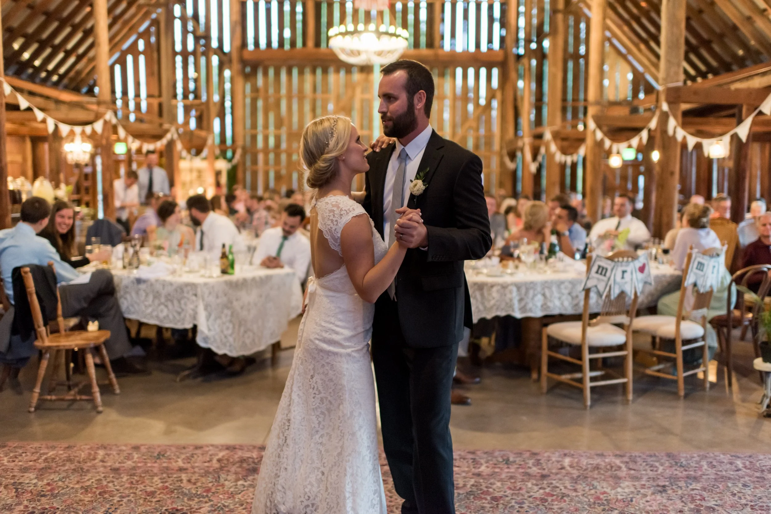 A bride and groom dancing at their wedding reception in a rustic wooden venue, with guest tables and wedding decor visible in the background.