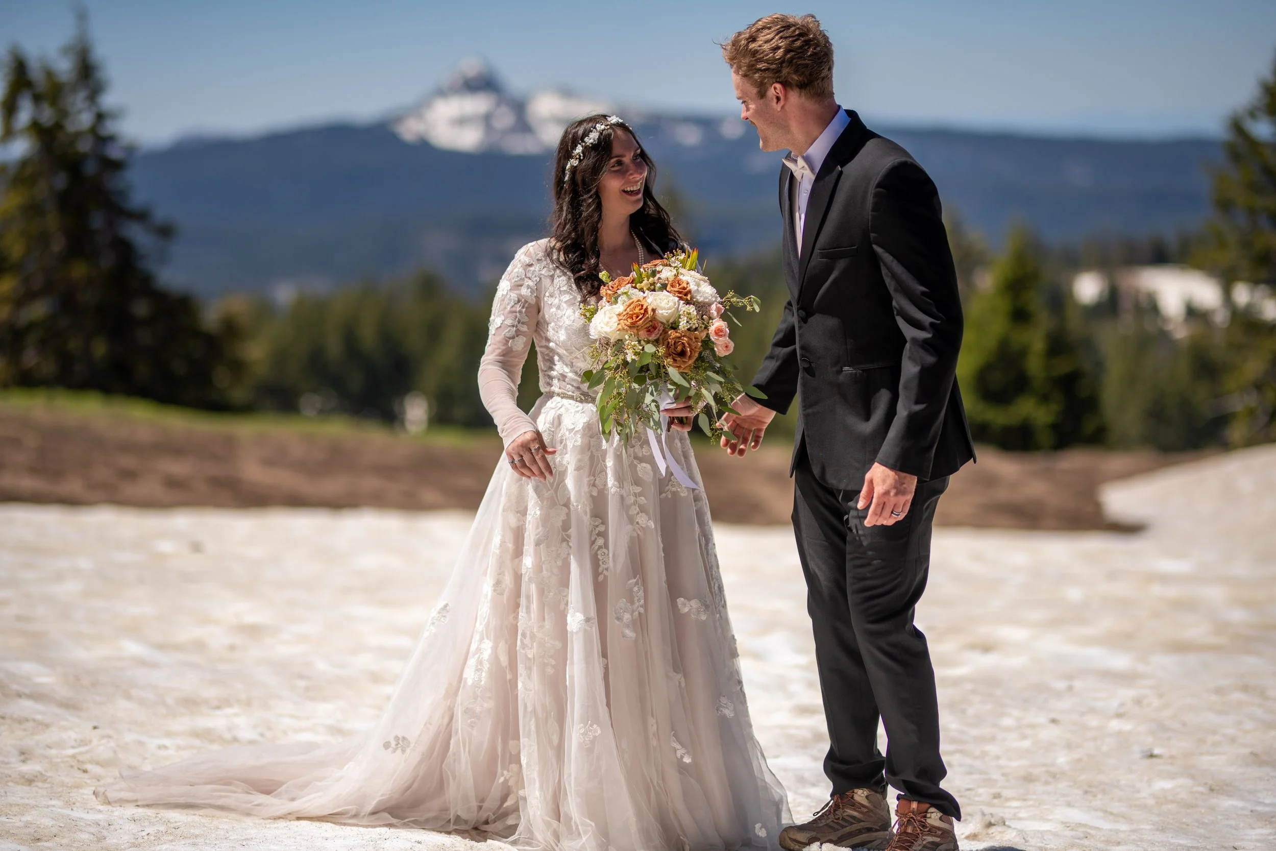 A bride and groom standing on a sandy outdoor location, holding hands, with a mountain in the background. The bride is wearing a white lace wedding dress and floral headband, holding a bouquet. The groom is dressed in a black suit with a white shirt,