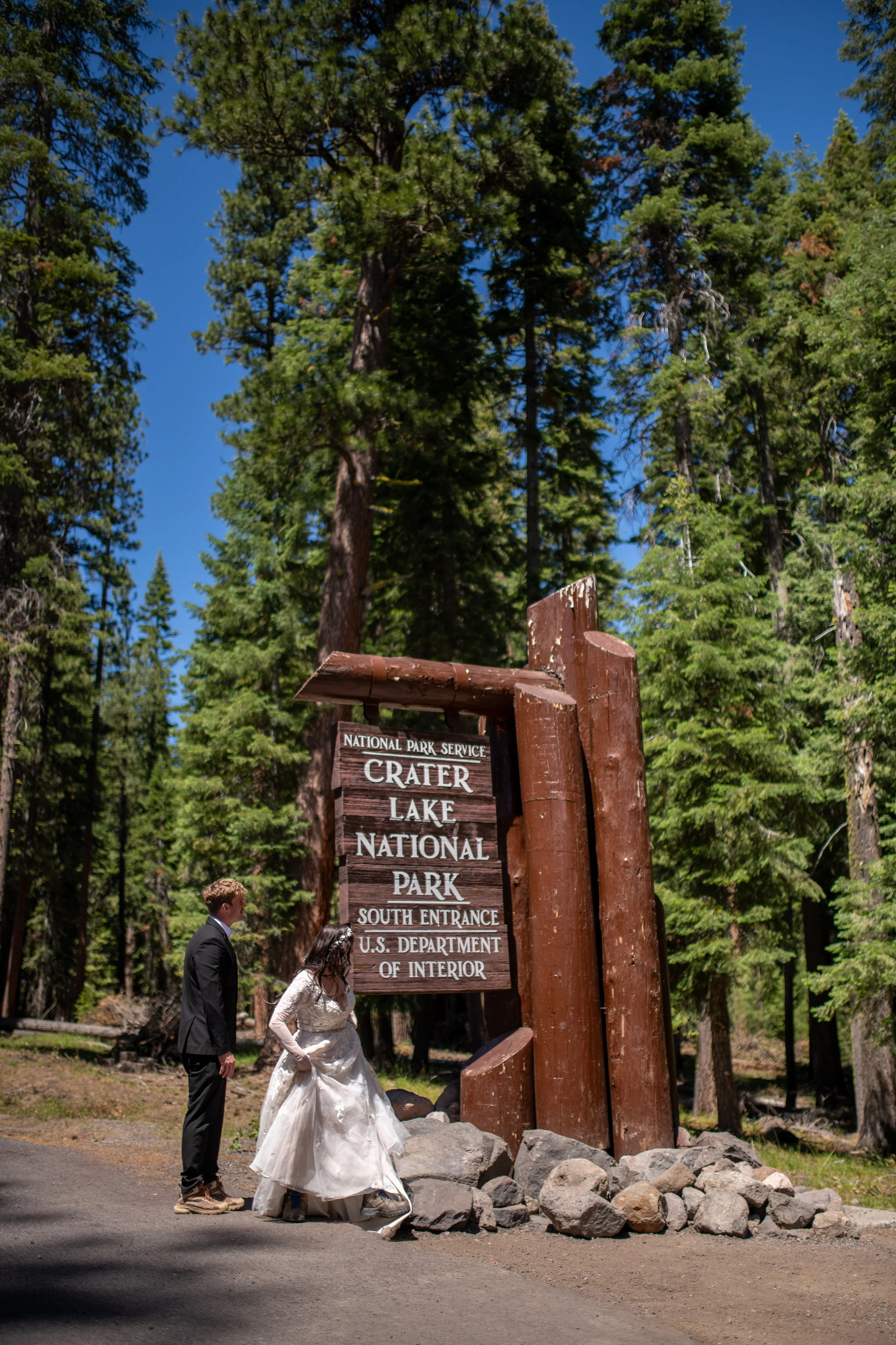A bride and groom walking past a large wooden sign at Crater Lake National Park, surrounded by tall pine trees and rocks.
