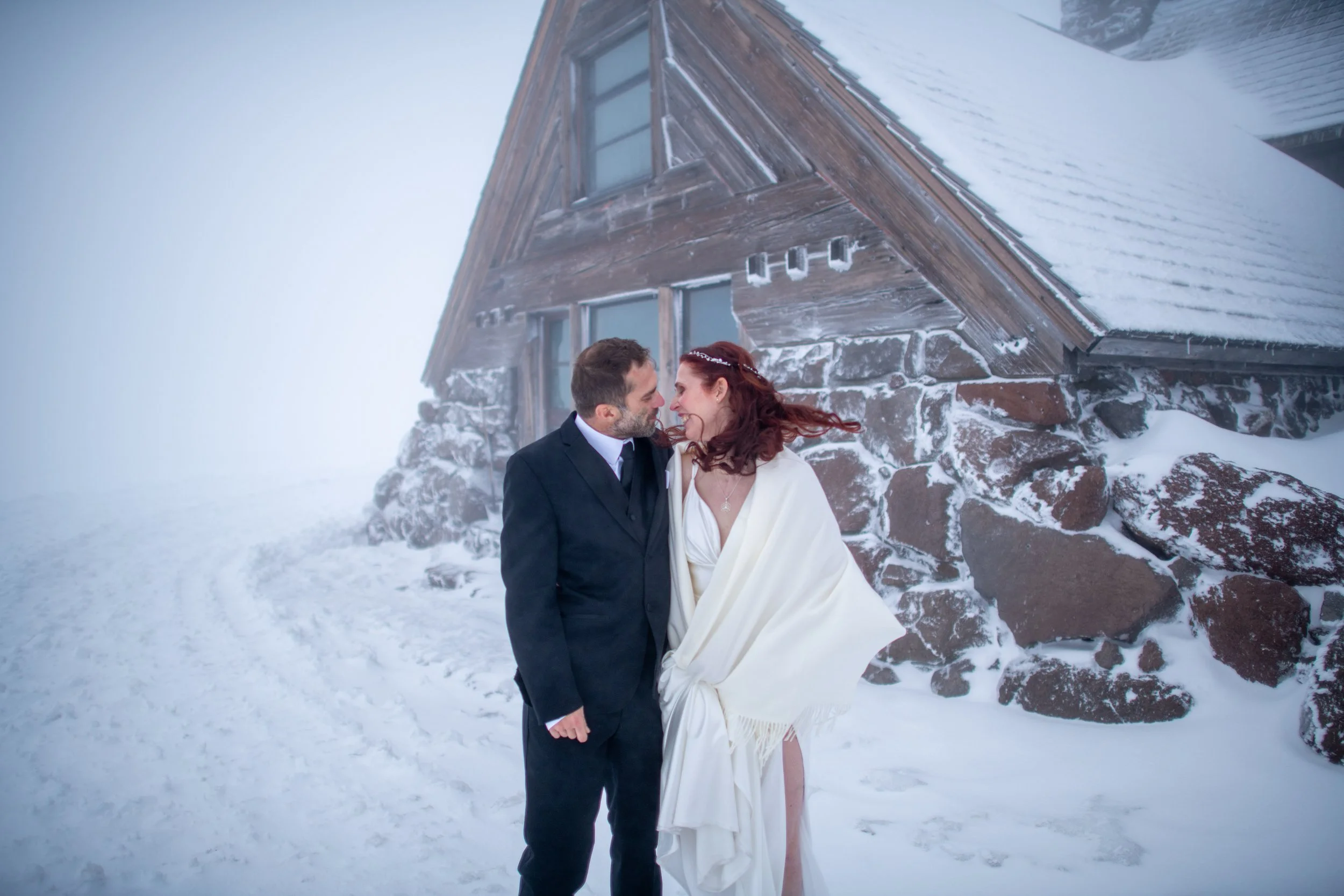 A couple dressed in wedding attire standing outdoors in a snowy mountainous area near a wooden cabin, with the groom in a black suit and the bride in a white dress and shawl, sharing a moment.