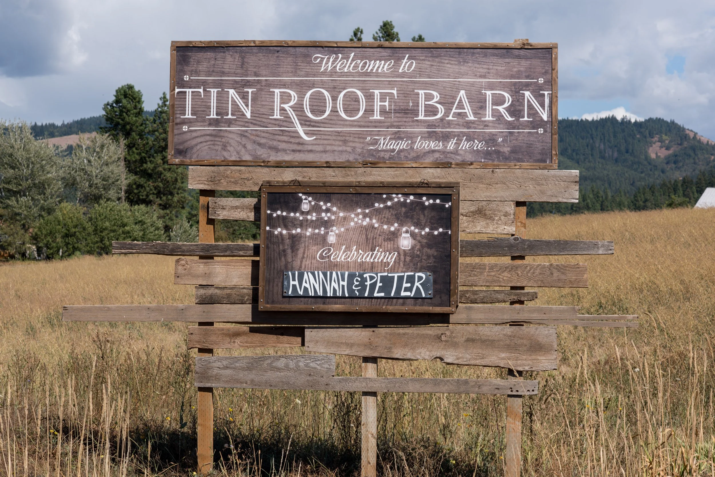 Wooden sign on a grassy field reading 'Welcome to Tin Roof Barn,' with the tagline 'Magic loves it here.' Below the main sign, there is a smaller decorative sign with string lights and the names 'Hannah & Peter.' In the background, there are trees, h
