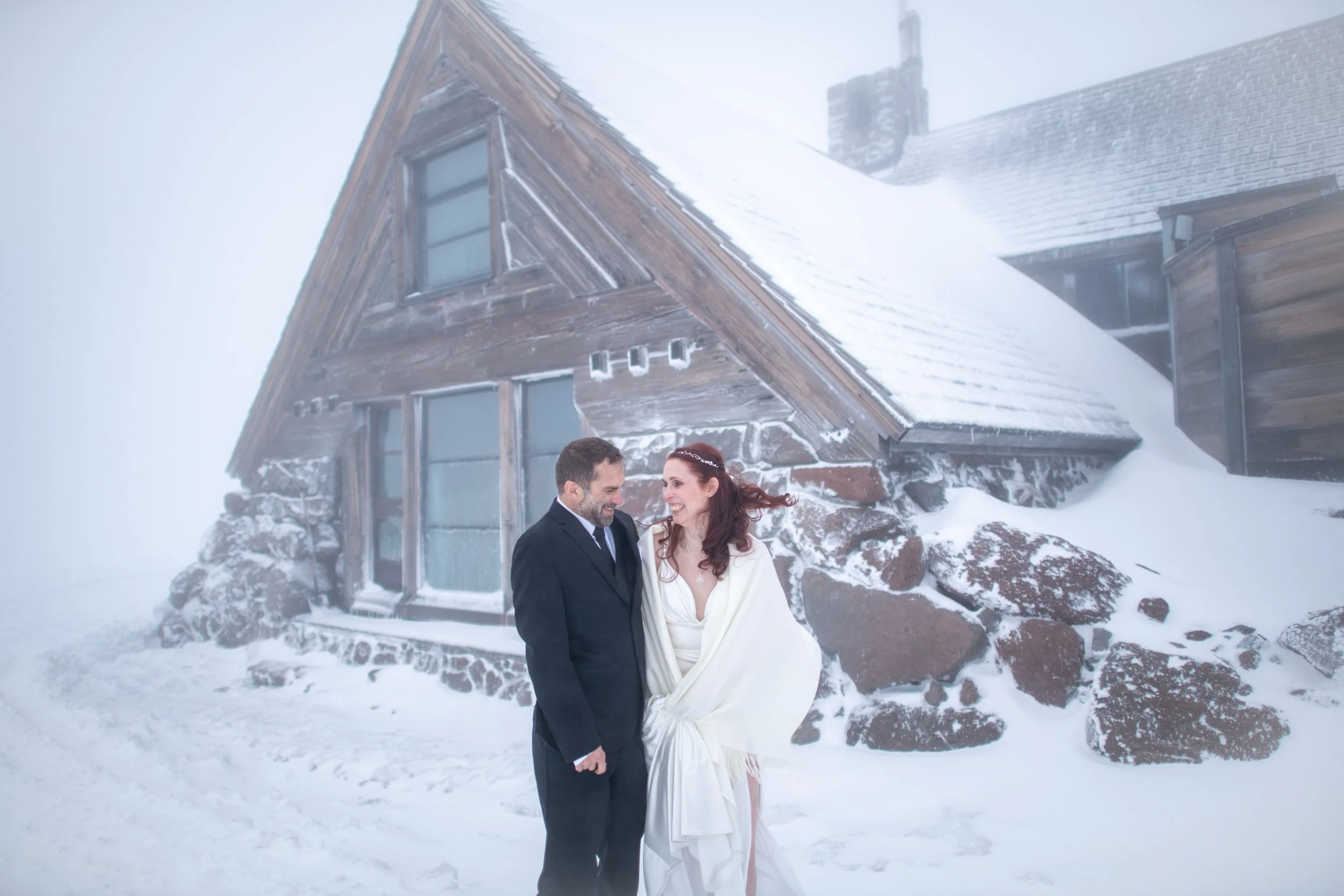 A newly married couple in wedding attire standing outside in snowy weather near a rustic wooden house with snow-covered roof.