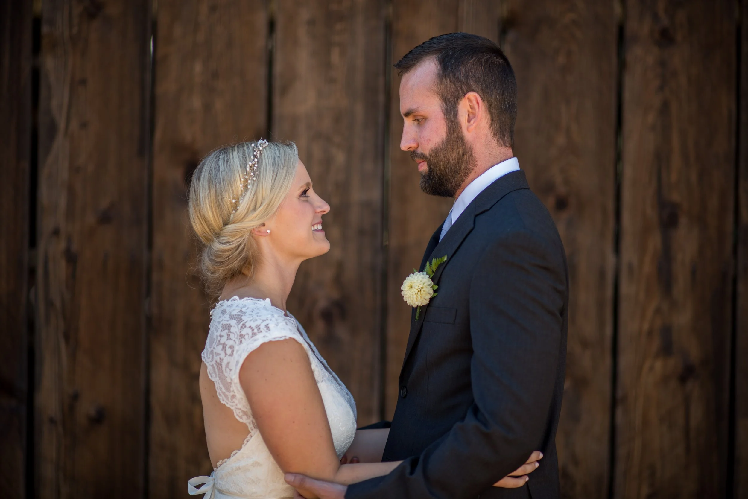 A bride and groom stand close together, looking into each other's eyes, in front of a wooden background. The bride has blonde hair and is wearing a white lace dress with a pearl headband. The groom has dark hair and a beard, and is dressed in a dark 