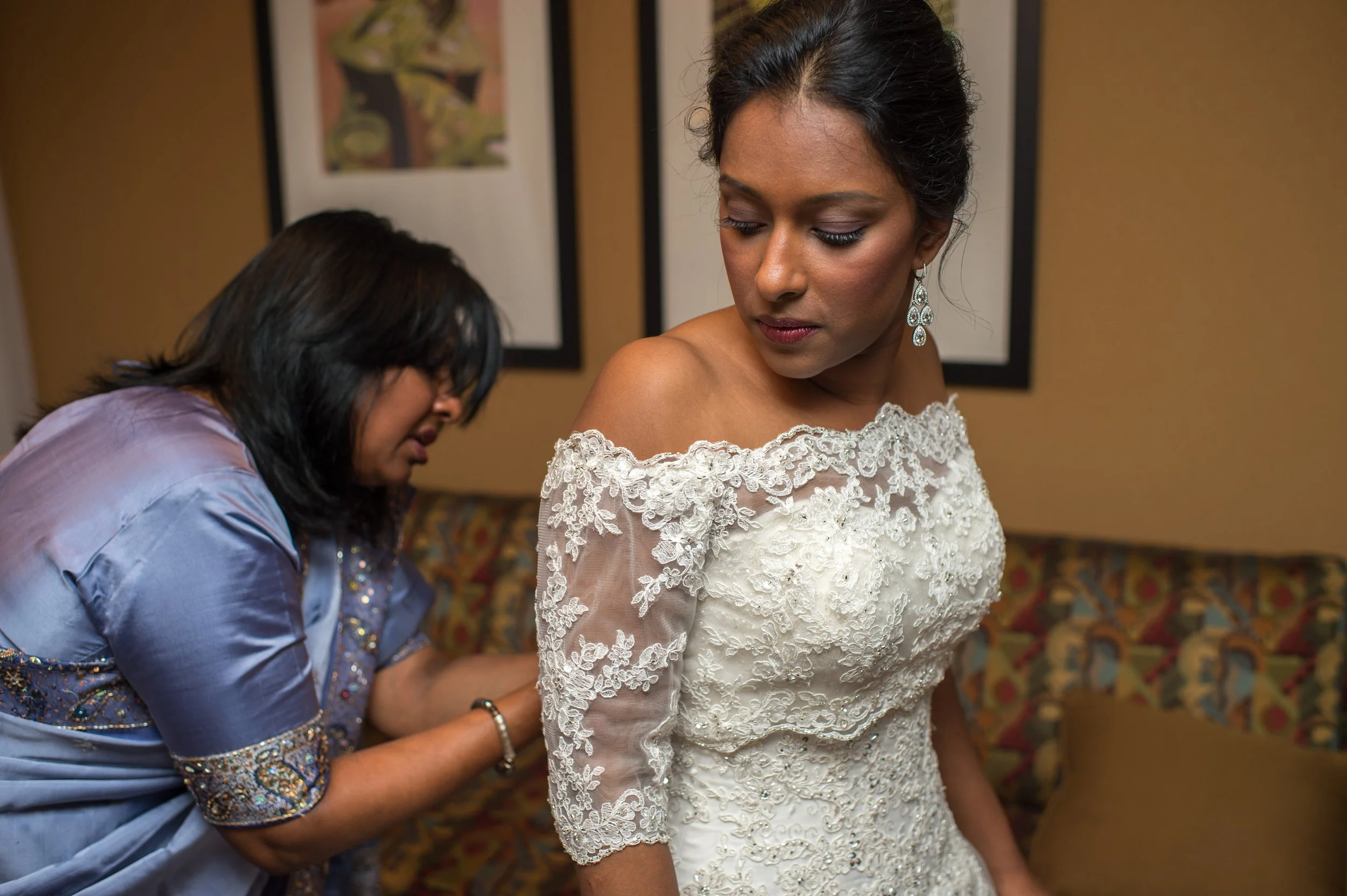 A woman in a wedding dress looks down as another woman helps her with her dress. The woman in the dress has earrings and hair styled up. The setting appears to be indoors with framed artwork in the background.