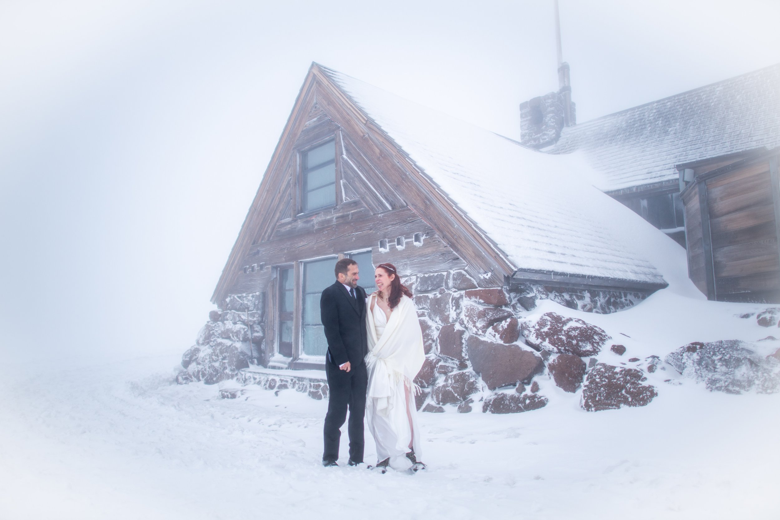 A couple in formal wedding attire standing outdoors in a snowy landscape with a rustic mountain cabin in the background.