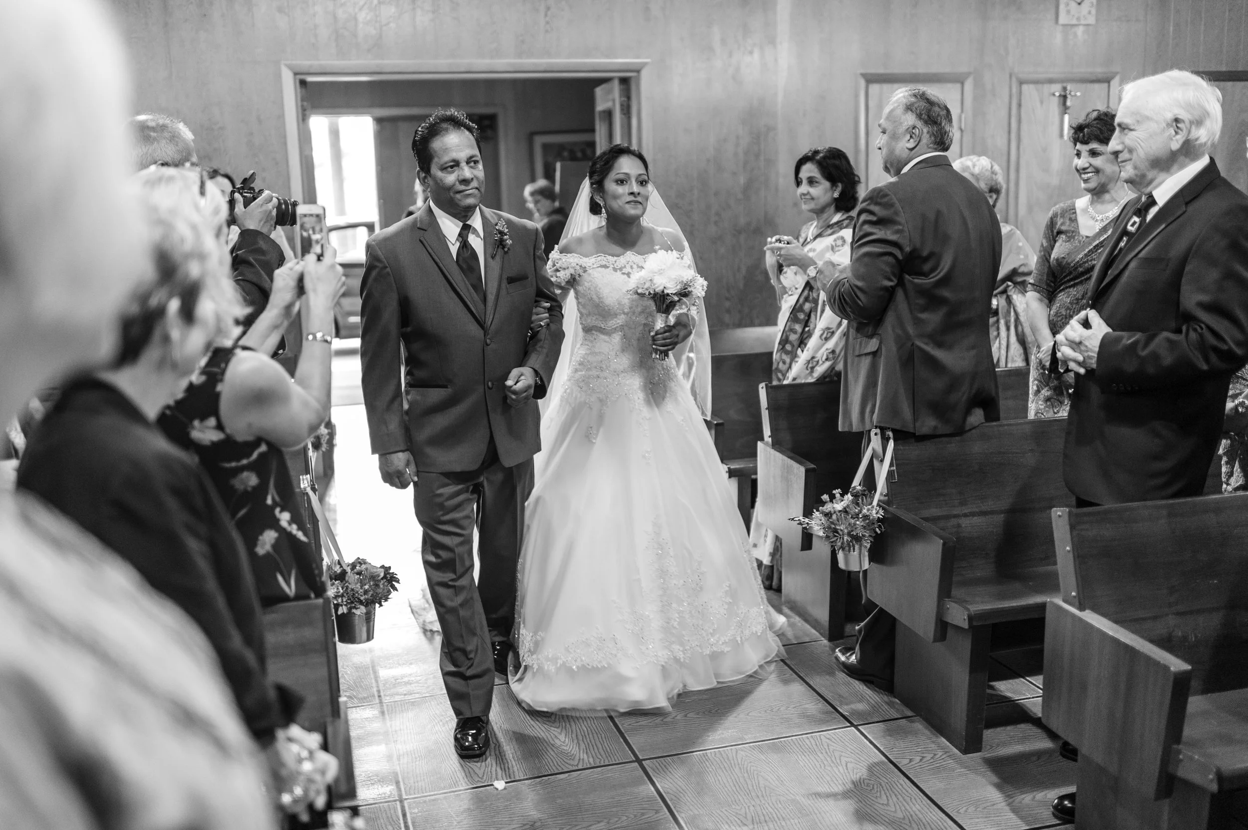 A bride in a wedding gown holding a bouquet walking down the aisle accompanied by a man in a suit at a wedding ceremony inside a church, with guests seated on pews and taking photos.