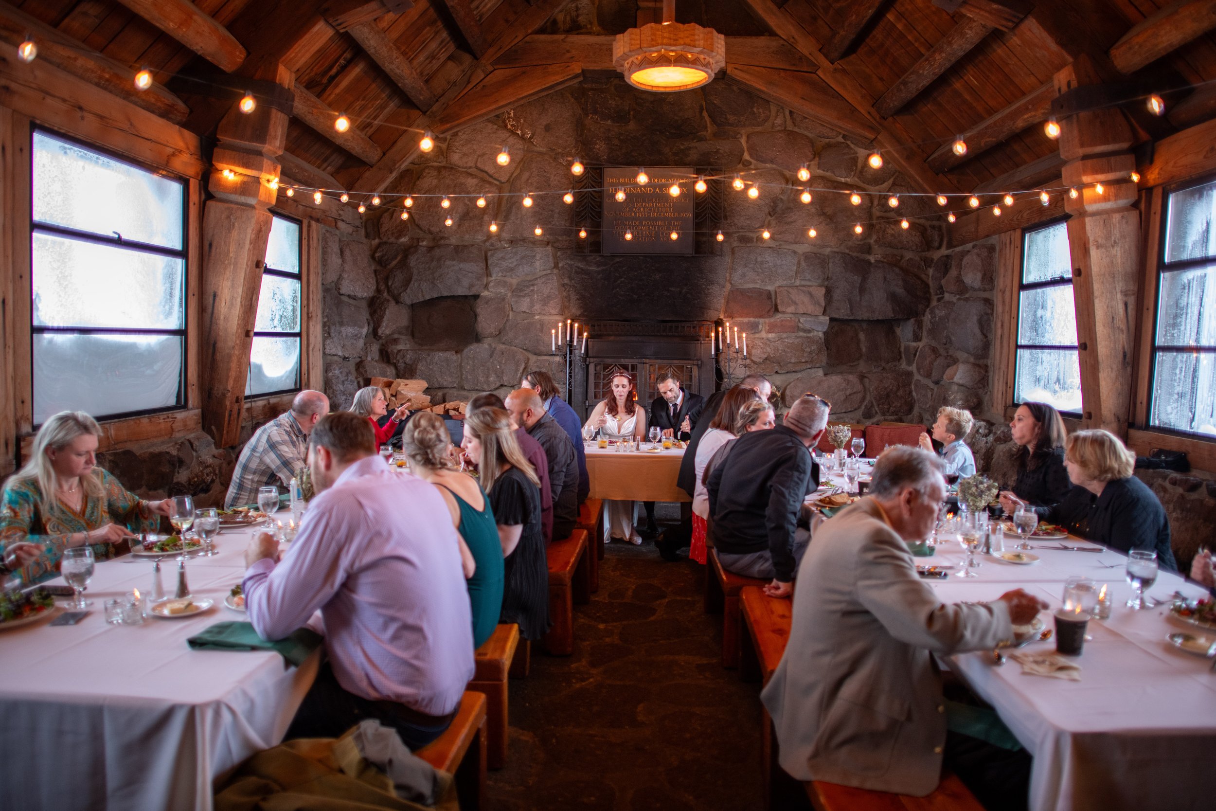 A group of people sitting at long tables during a meal in a rustic, stone-walled dining room with string lights hanging from the wooden ceiling.
