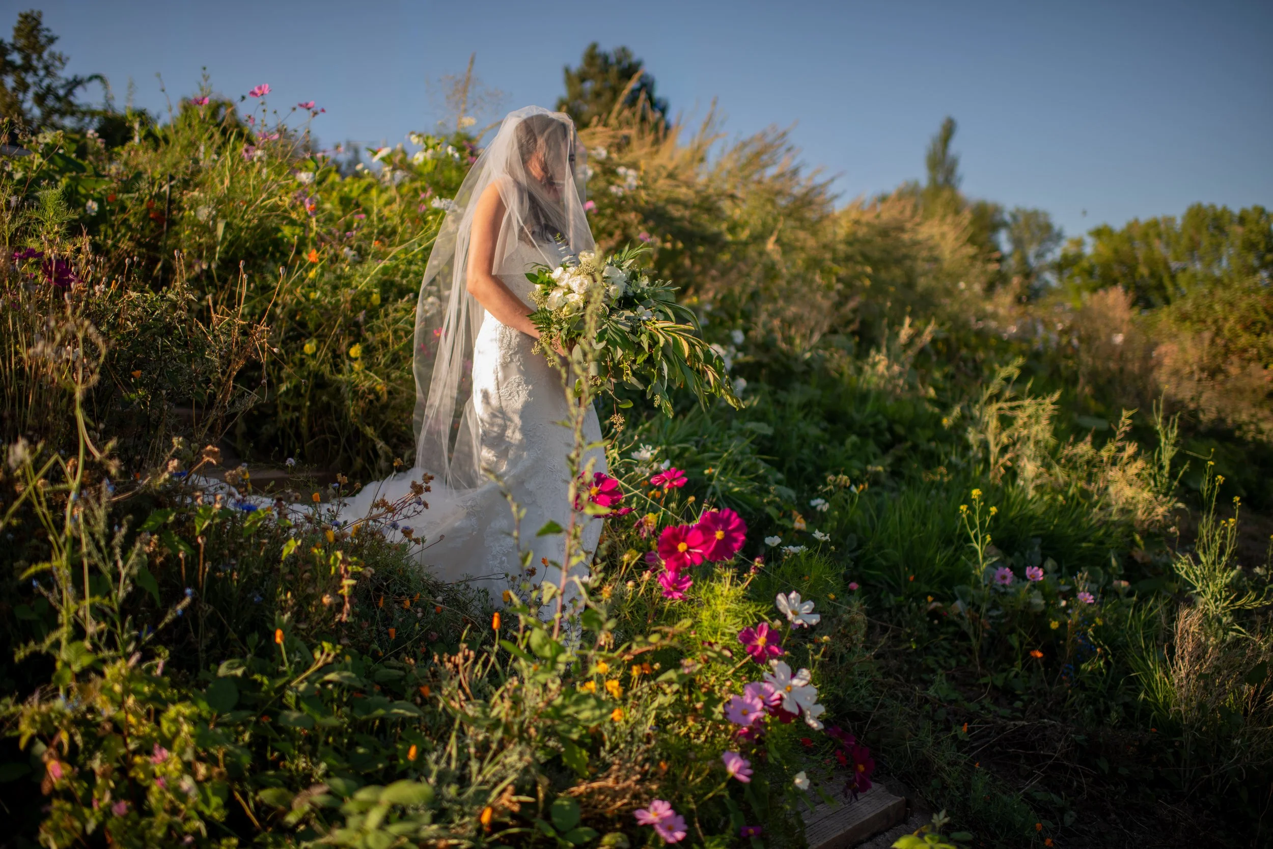A woman in a wedding dress with a veil holding a large bouquet of white flowers, standing among colorful flowers and greenery outdoors on a sunny day.