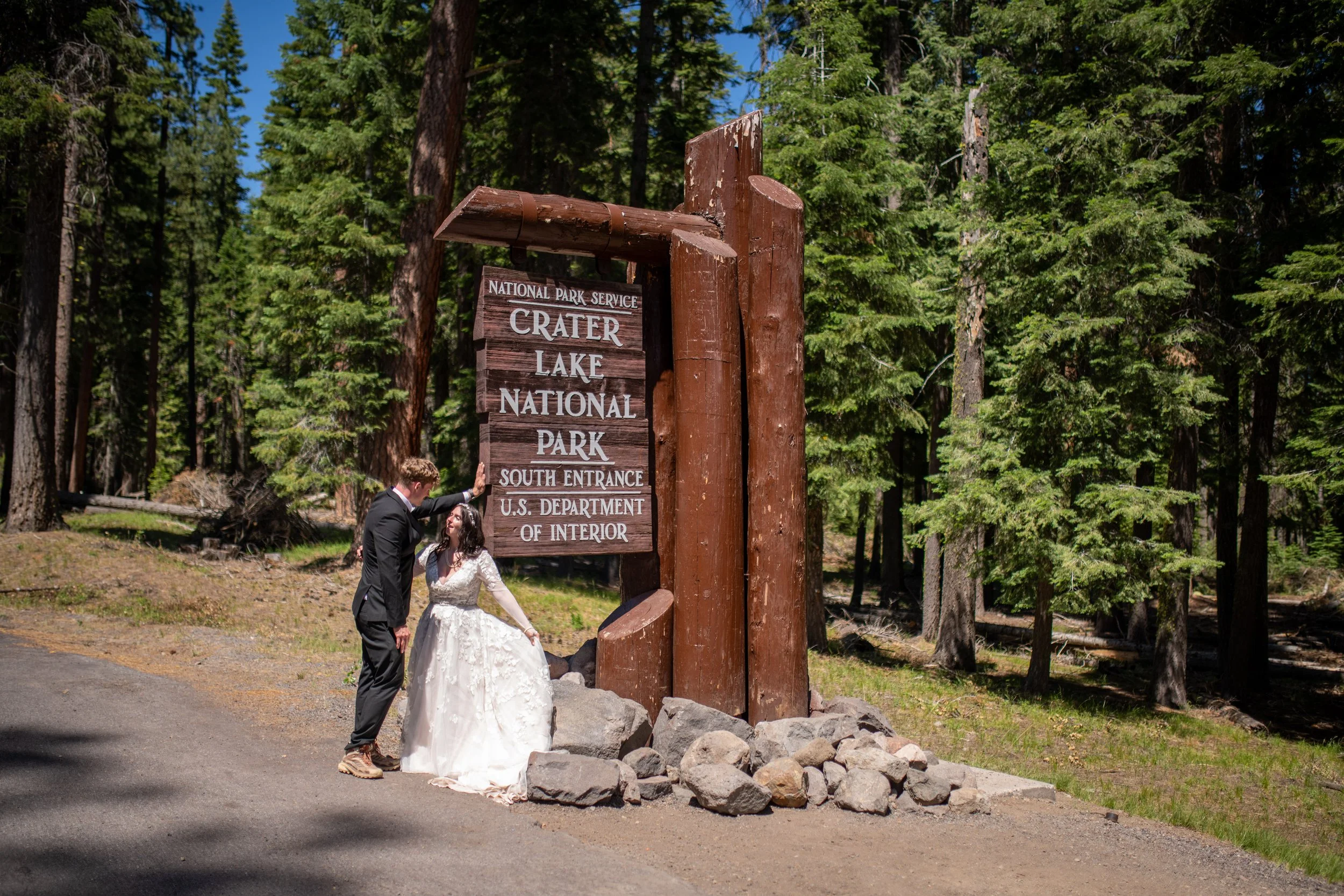 A couple in wedding attire standing near a wooden sign that reads 'Crater Lake National Park South Entrance'; surrounded by tall green trees in a forested area.