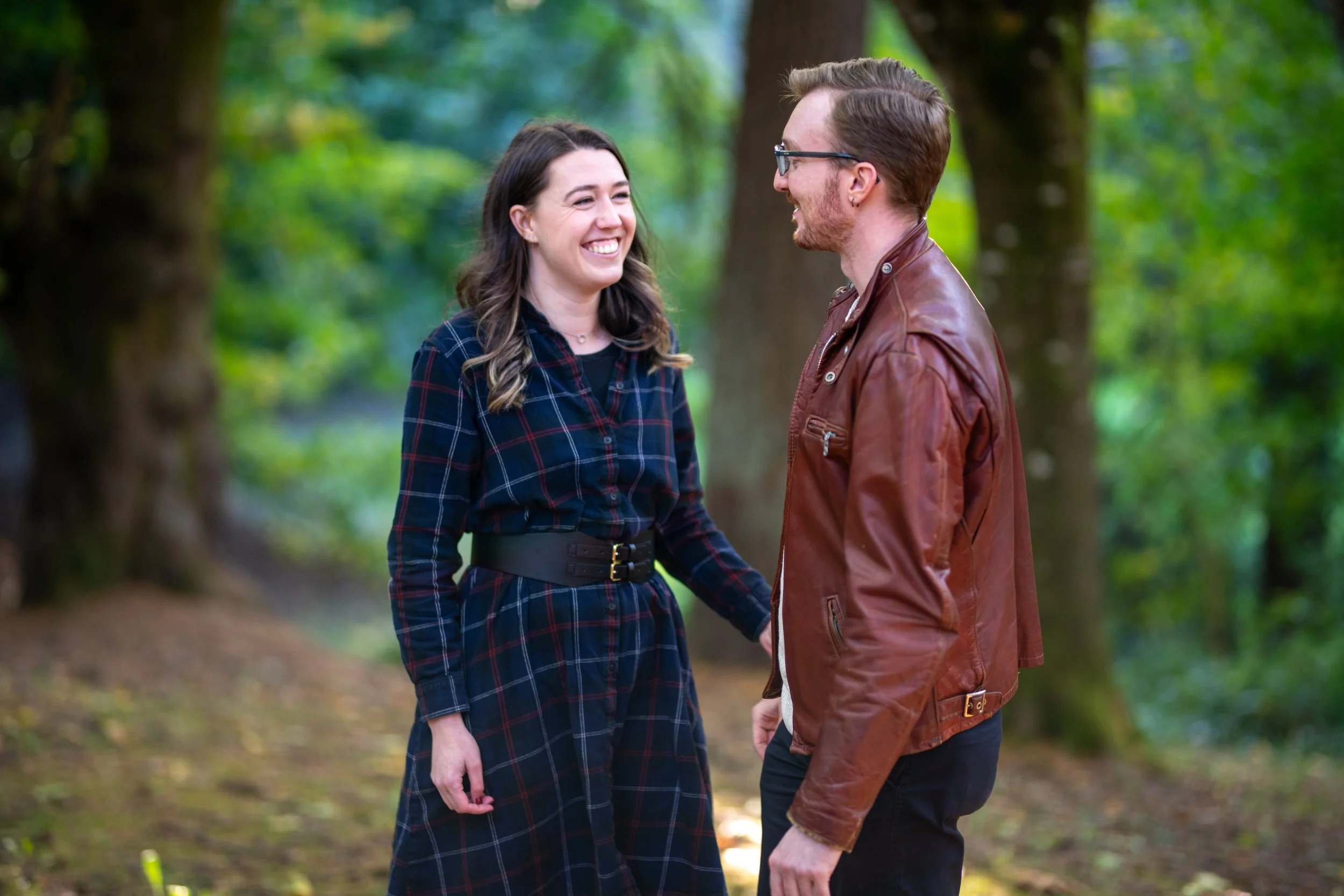 A man and woman smiling and holding hands, standing face to face outdoors in a wooded area with trees and greenery in the background.