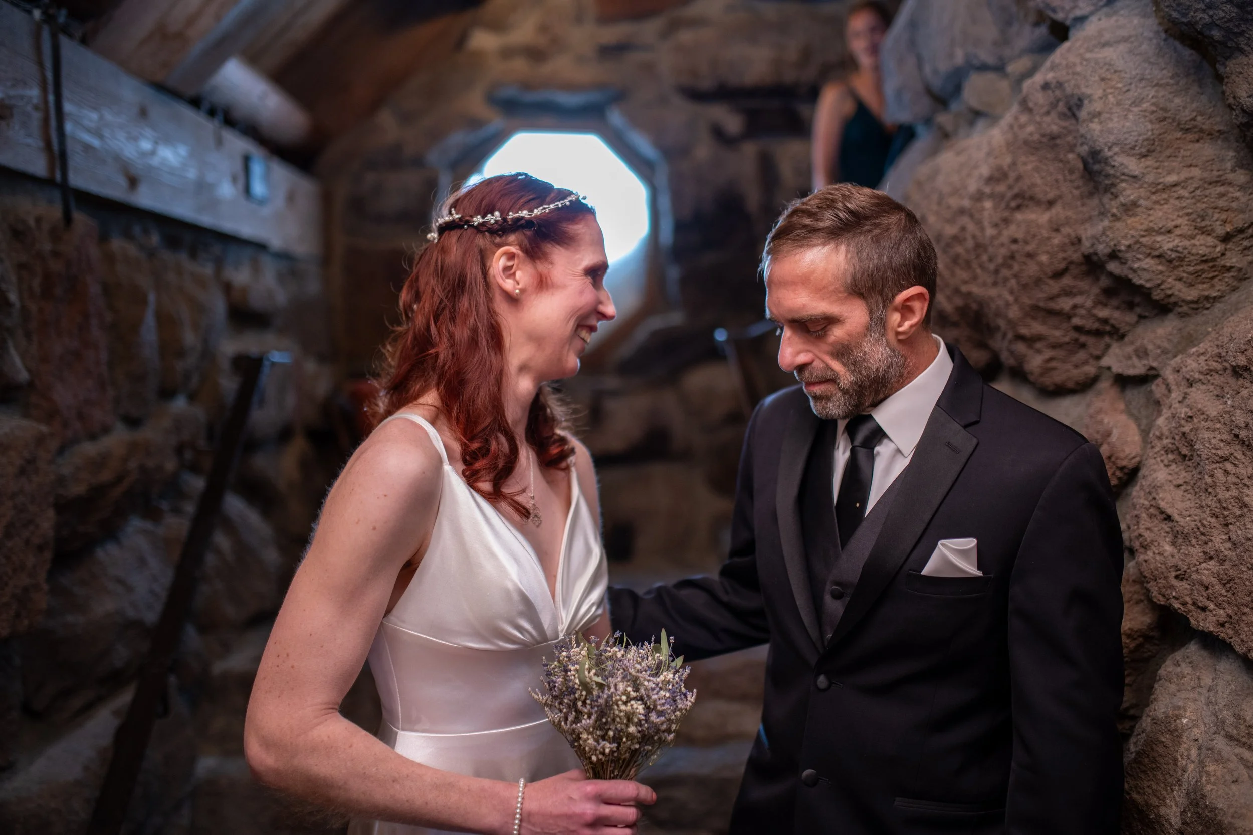 A bride and groom sharing a moment during their wedding ceremony in a rustic stone altar setting. The bride has red hair and is wearing a white wedding dress with a floral headband, holding a bouquet. The groom is in a black suit with a white shirt a