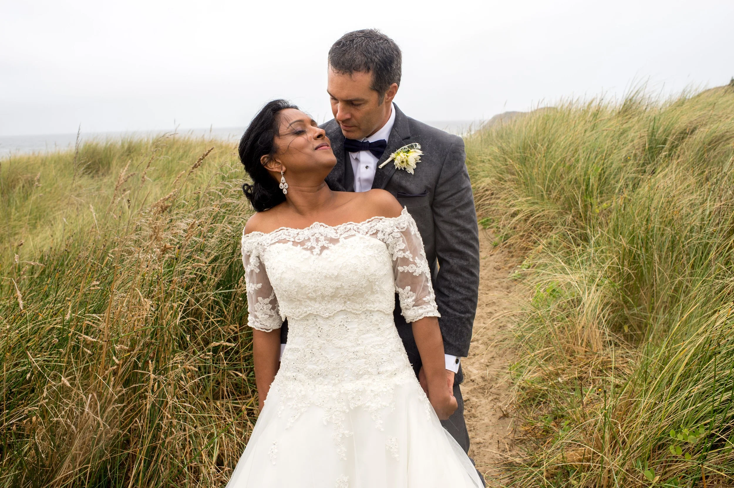 A bride and groom are standing on a sandy path surrounded by tall grass, with the ocean in the background, during daytime. The bride is in a white lace wedding dress with off-the-shoulder sleeves, and the groom is in a gray suit with a bowtie. They a