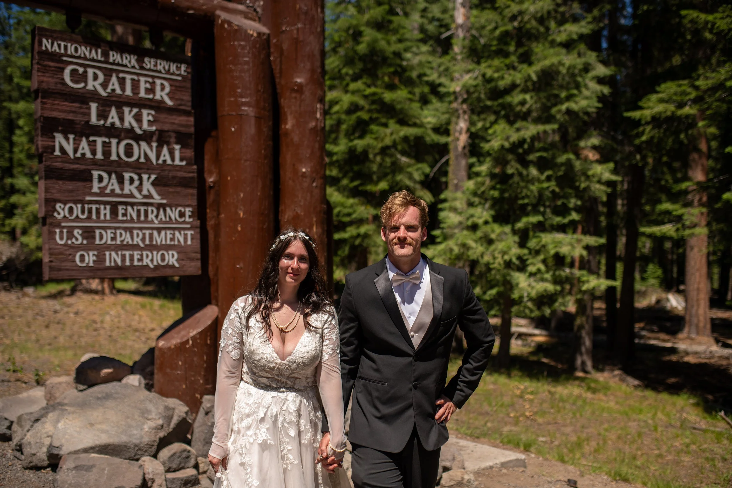 A bride and groom standing outside near a wooden sign that reads 'Crater Lake National Park South Entrance' surrounded by trees.