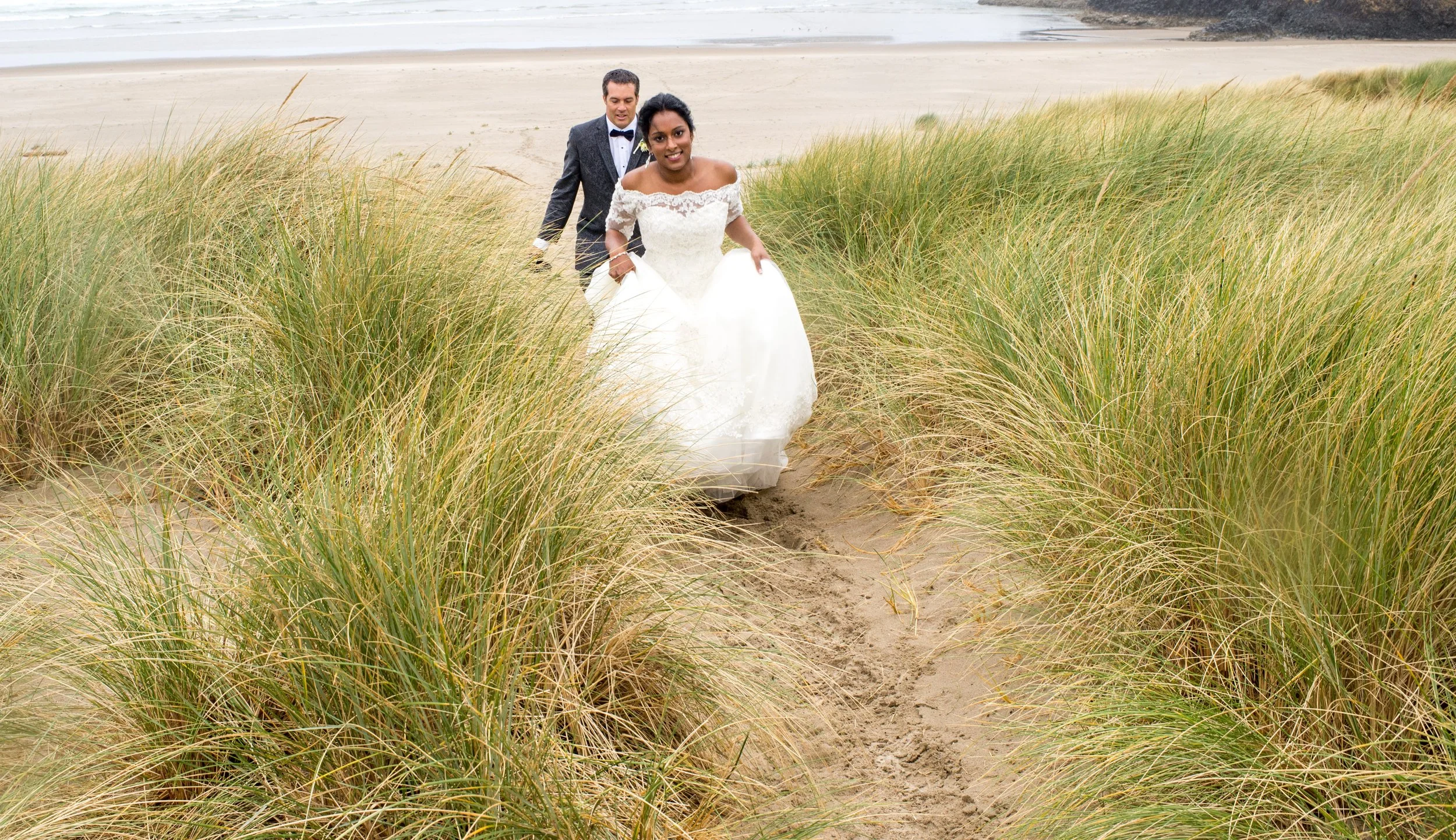 A bride and groom walking on a sandy beach through grassy dunes, with the ocean in the background.